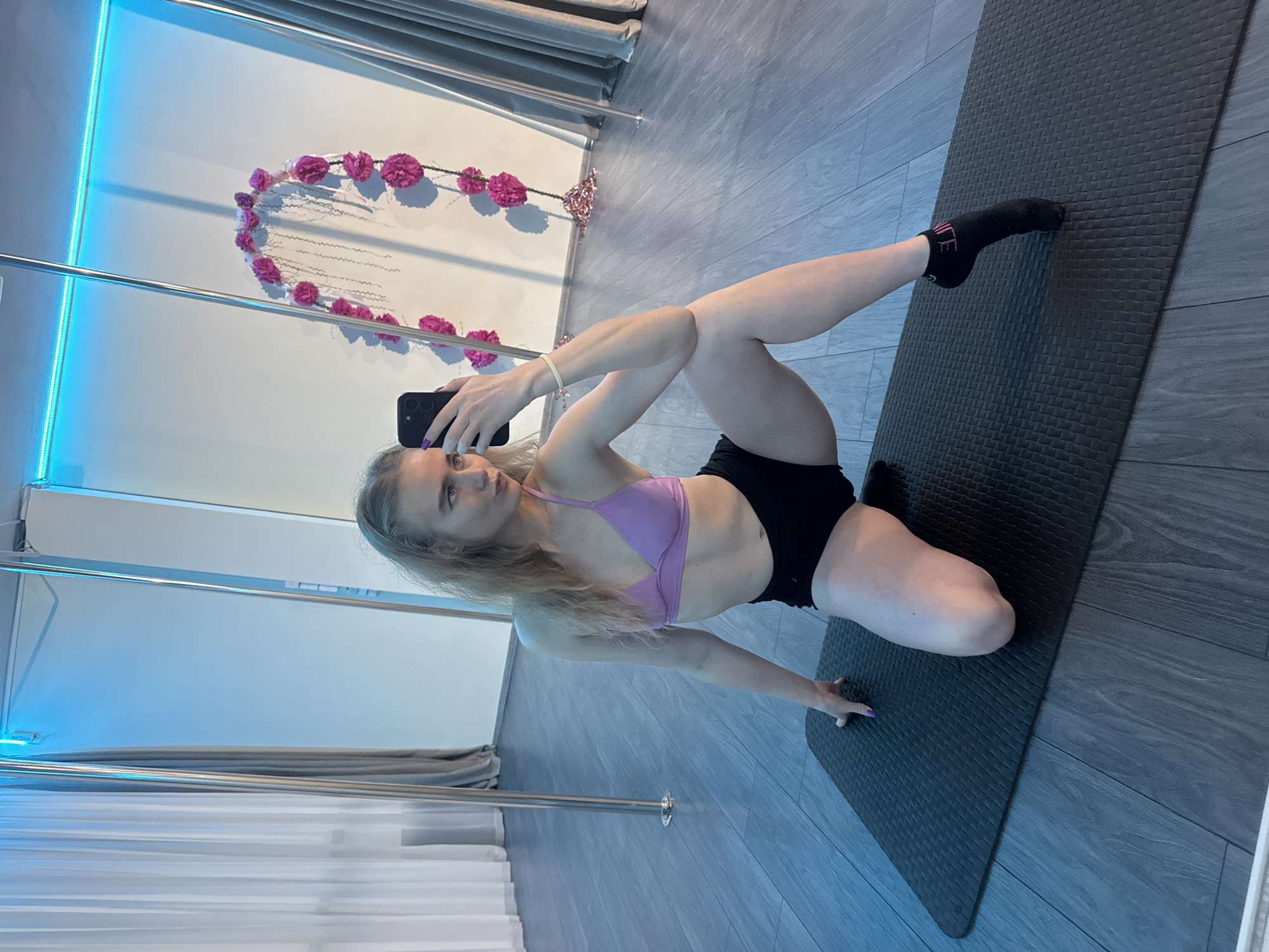 A woman with blonde hair, wearing a light purple sports bra and black shorts, is taking a selfie with her black smartphone while performing a stretch on a dark gray exercise mat. She is kneeling with her right knee on the mat, right foot in a black sock, while her left leg is bent behind her. The backdrop includes a dance pole studio with a chrome dance pole, a white wall adorned with a heart-shaped floral decoration, and gray curtains. The studio features blue lighting. The location and city are unknown.