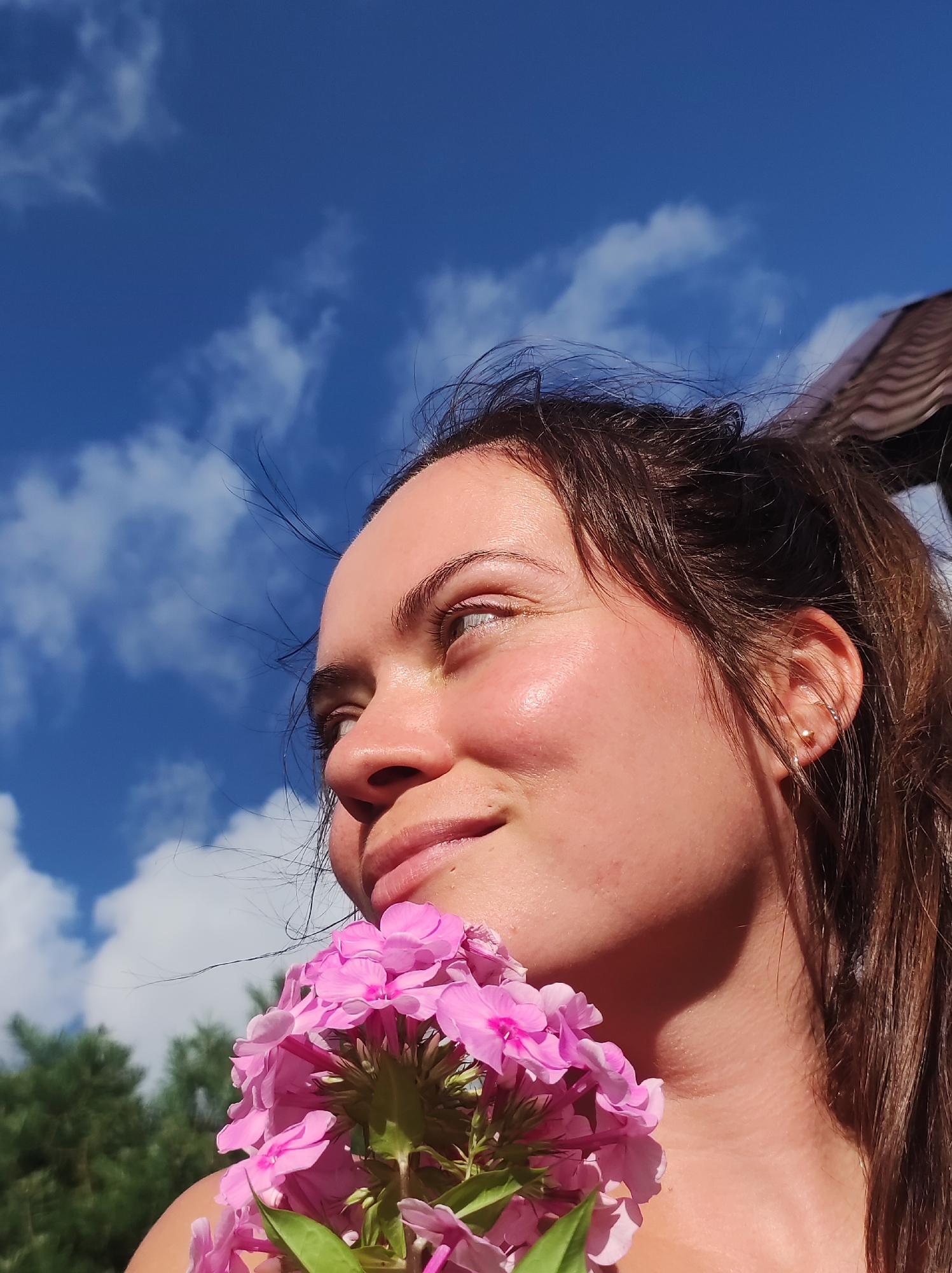 The image is a close-up, low-angle shot of a young woman's face, captured outdoors against a bright blue sky with scattered white clouds. She is looking upwards and to her left, with her head tilted slightly. Her hair is dark brown and pulled back into a ponytail. She is holding a cluster of bright pink flowers near her chin. Her skin appears smooth and slightly flushed, with sunlight highlighting her cheekbones. A portion of a brown, ridged structure, possibly a roof or awning, is visible in the upper right corner of the frame. Green foliage, likely trees, forms a blurred background at the bottom. The scene is illuminated by natural sunlight, creating soft shadows on her face. No specific location in the city of Фоки, Russia, is identifiable from the image itself.