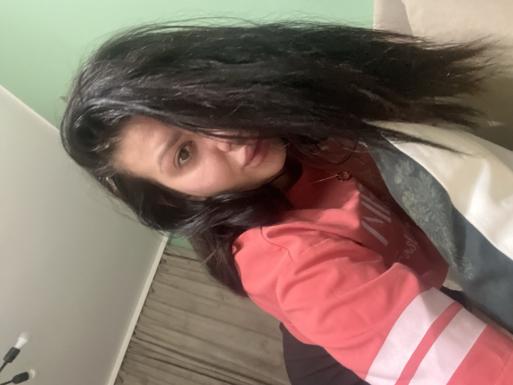 A young woman with long, dark hair is lying down on a couch, her face partially obscured by her hair. She is wearing a pink long-sleeved shirt with white stripes on the sleeves and a necklace with a circular pendant. The background consists of a light green wall, brown curtains, and a portion of a white duvet. There are also light fixtures visible on the ceiling. The image was taken indoors, likely in a residential setting, and no specific Plovdiv landmarks are visible.