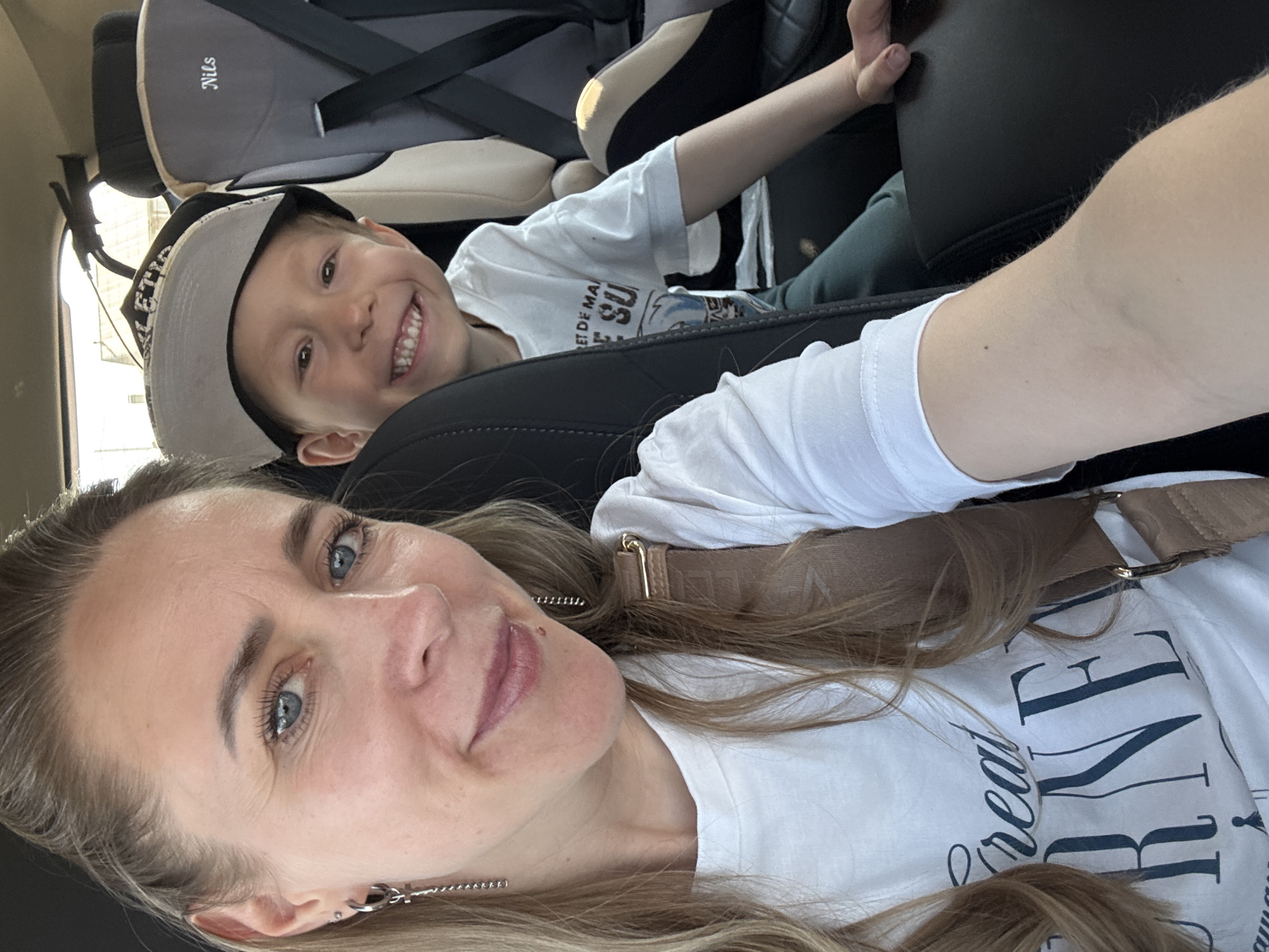 A selfie taken inside a vehicle in Chaikovsky, Russia, features a woman and a young boy. The woman, with fair skin, blue eyes, and light brown hair, is in the foreground. She wears a white t-shirt with dark lettering and a brown shoulder strap. The boy, with light skin, brown eyes, and a baseball cap, is in the background, smiling and positioned in a child's car seat. The interior of the car includes black leather seats and a gray child's car seat.