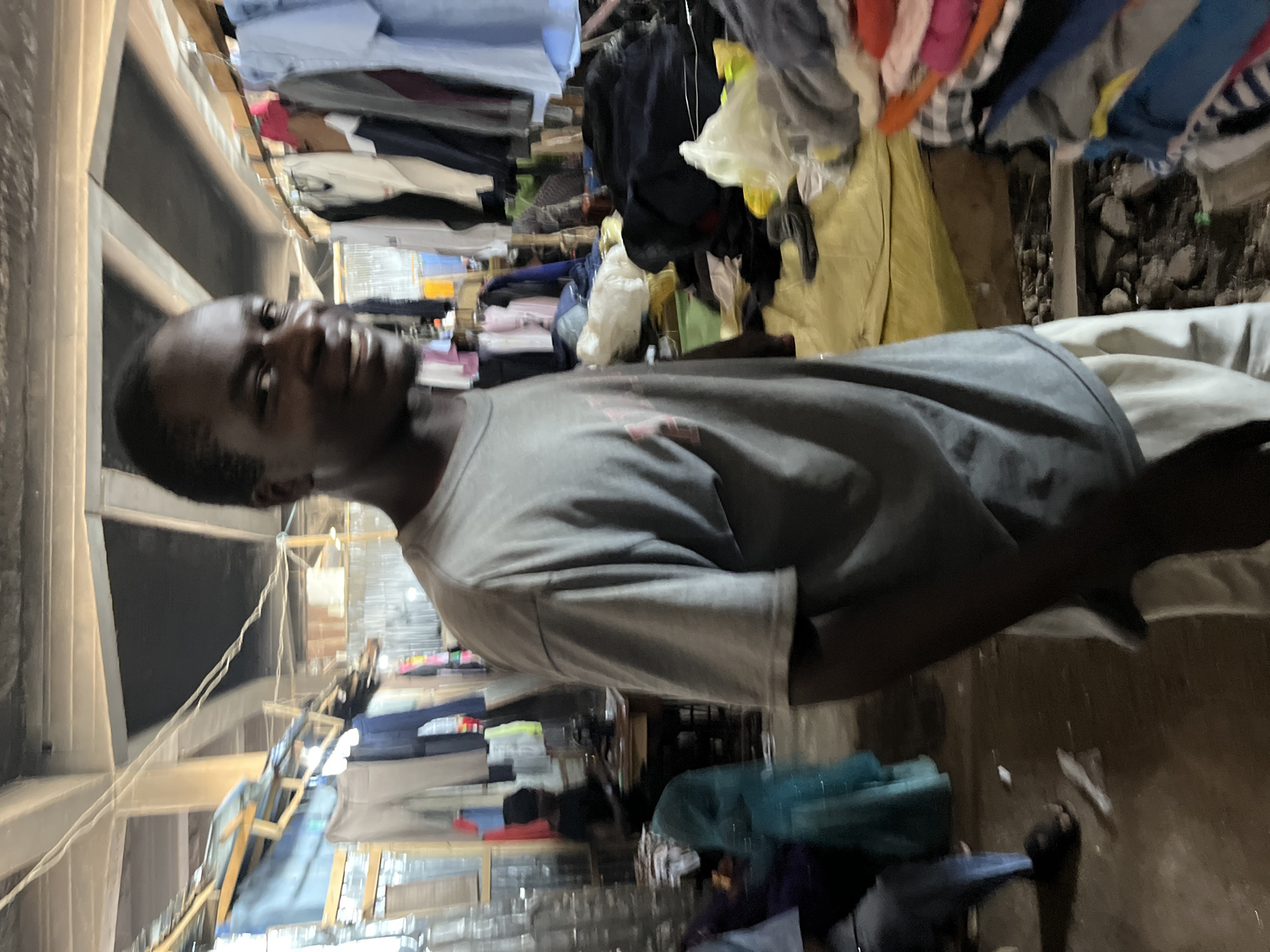 The image depicts a person in what appears to be an informal market or shop, potentially in Jos, Nigeria. The person, an adult, is wearing a grey t-shirt. The background is filled with hanging clothes and other textiles, creating a cluttered and cramped environment. Some items are stacked overhead, while others are displayed on makeshift shelves. The floor is visible, showing a worn surface.