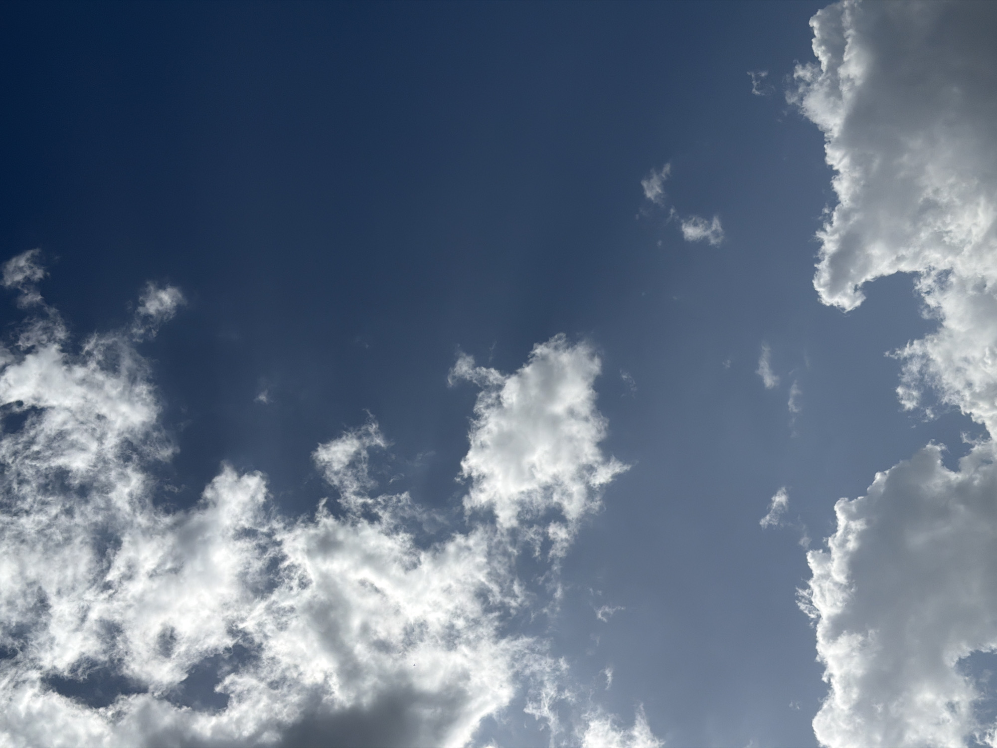 The image depicts a daytime sky with scattered clouds. The sky ranges in color from a deep blue in the upper left to a lighter blue/gray towards the right. Clouds are concentrated along the lower left and right edges, as well as a patch in the center. The clouds vary in density, with some appearing wispy and translucent, and others are dense and opaque. No city or people are visible in the image, so location is undetermined.