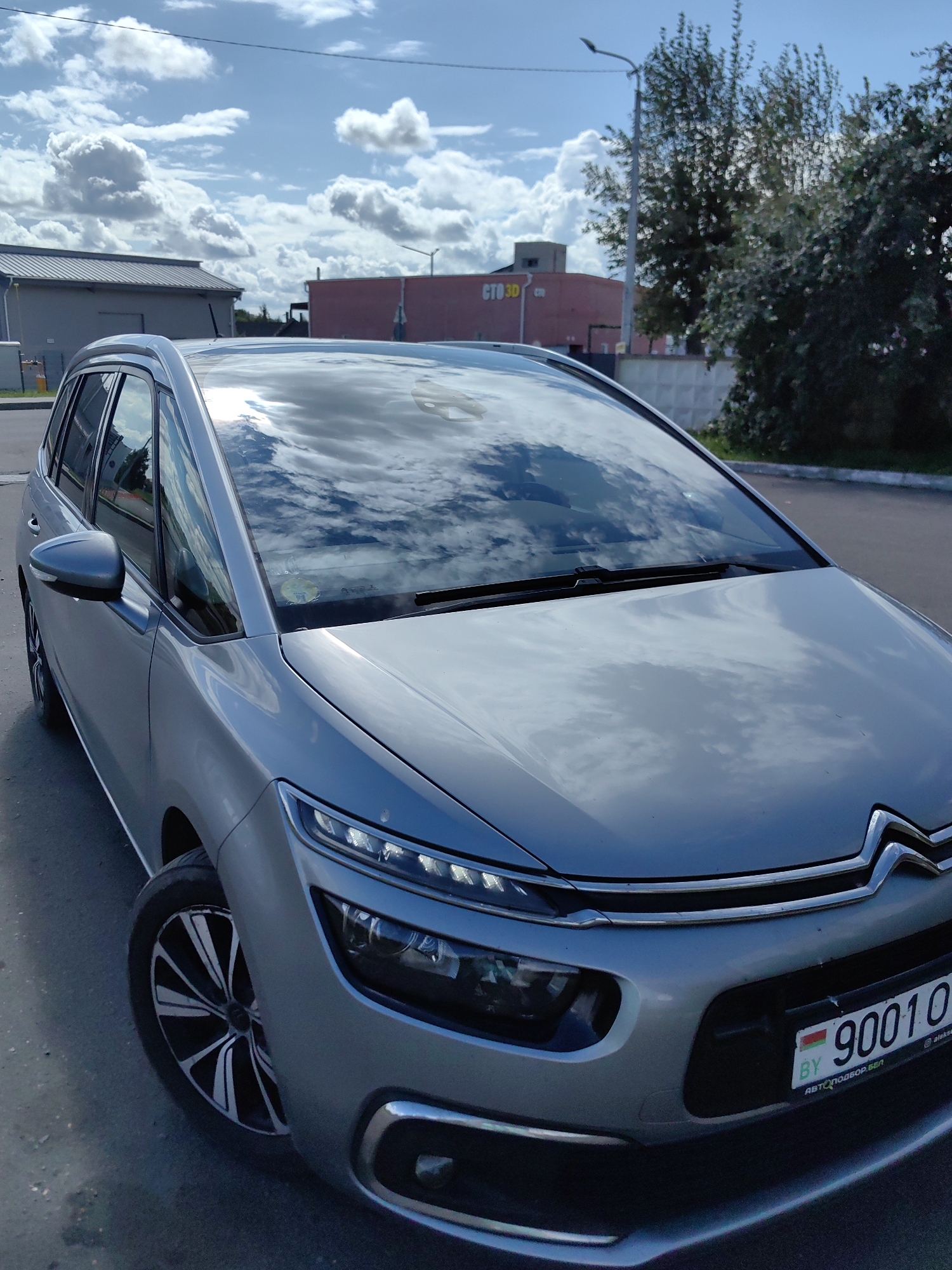 A silver Citroën C4 Picasso is parked on an asphalt surface. The car is a multi-purpose vehicle with a distinctive front end featuring LED headlights and a chrome grille. The license plate is visible and reads "BY 9001 OA". In the background, there are buildings, trees, a street lamp, and a cloudy sky. The image appears to have been taken outdoors during the day. The building on the left is gray and appears to be a commercial or industrial structure. The building in the center-left is a pink, rectangular structure with the text "СТО 3D" on its facade. The sky is blue with scattered white clouds. The overall scene suggests an urban or suburban environment.
