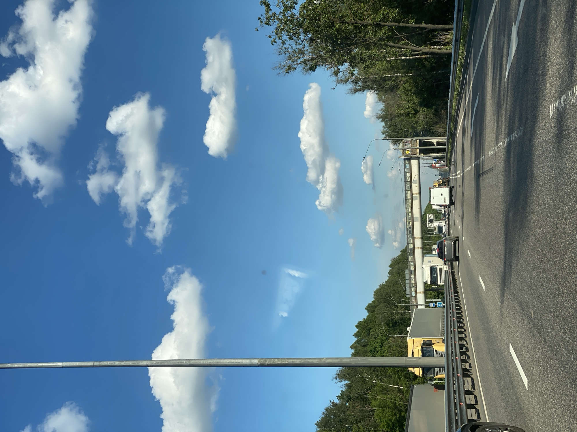 This image, taken from a moving vehicle, captures a daytime view of a multi-lane highway. The sky is a vibrant blue with scattered, fluffy white cumulus clouds.
In the foreground on the left, a tall, grey metal pole rises vertically, partially obscuring the sky. The asphalt road surface, prominent in the lower half and extending towards the right, features crisp white lane markings, including solid lines, dashed lines, and a large white arrow indicating direction of travel. A grey metal guardrail separates the highway lanes on the right side.
Vehicles are traveling on the highway; a dark grey SUV is visible in the rightmost lane, followed by a white truck. Further ahead and to the left, a yellow truck with a grey trailer is partially seen. Other vehicles, including more trucks, are in the mid-distance. The highway is flanked by dense green foliage, primarily deciduous trees, on both sides.
In the mid-ground, an overhead gantry or bridge structure spans across the highway. The scene is brightly lit by natural sunlight, casting subtle shadows on the road, suggesting a clear and sunny day. No legible text is discernible on any signs or vehicles within the image.