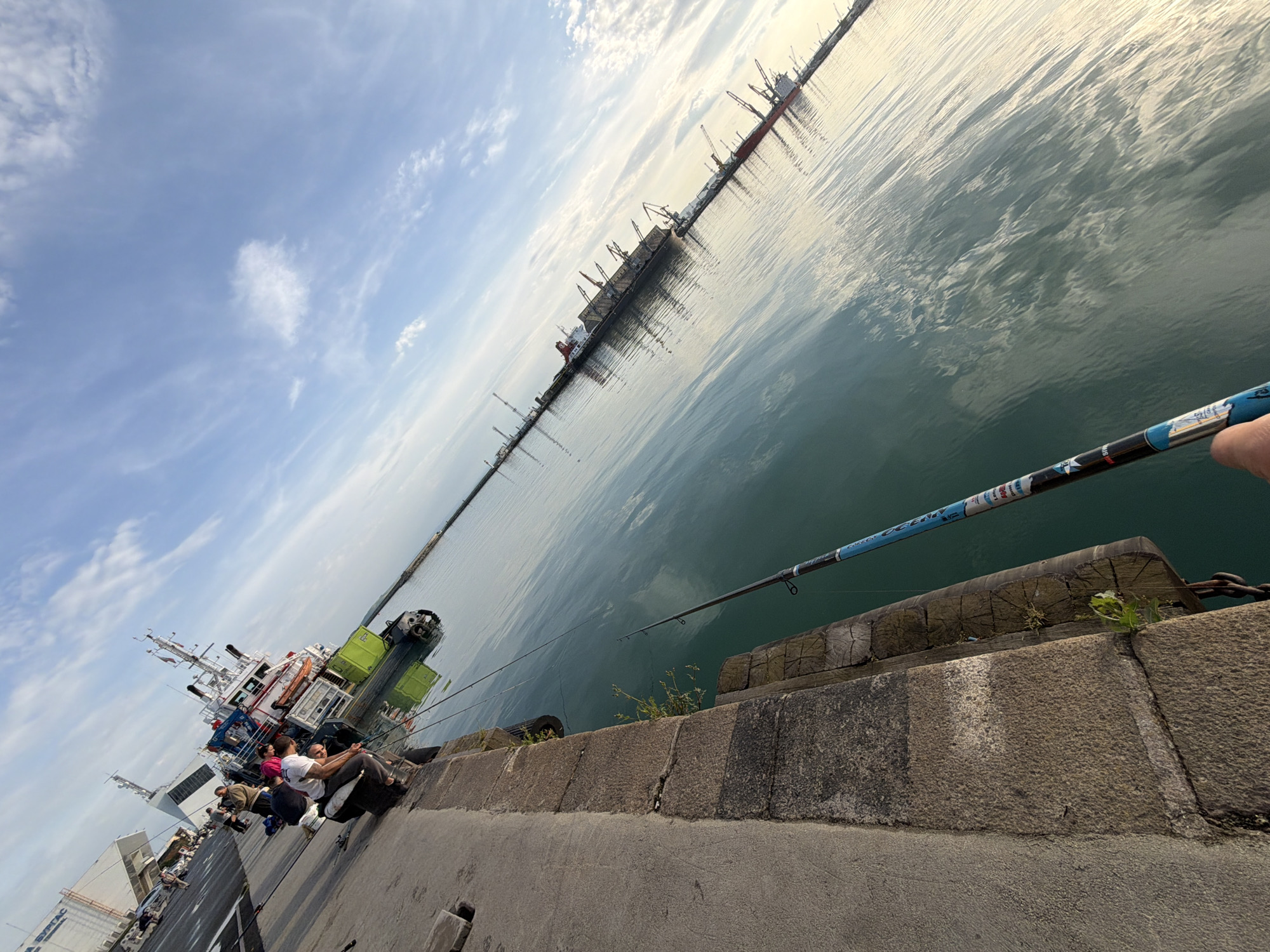 The image depicts a pier in Burgas, Bulgaria. In the foreground, a concrete embankment leads to a stone barrier alongside the water. A blue fishing rod extends over the barrier towards the sea. Several people are sitting on the embankment, engaged in fishing. A large ship is docked nearby, and in the distance, there are more cargo ships and cranes visible along the coast. The sky is partly cloudy.
