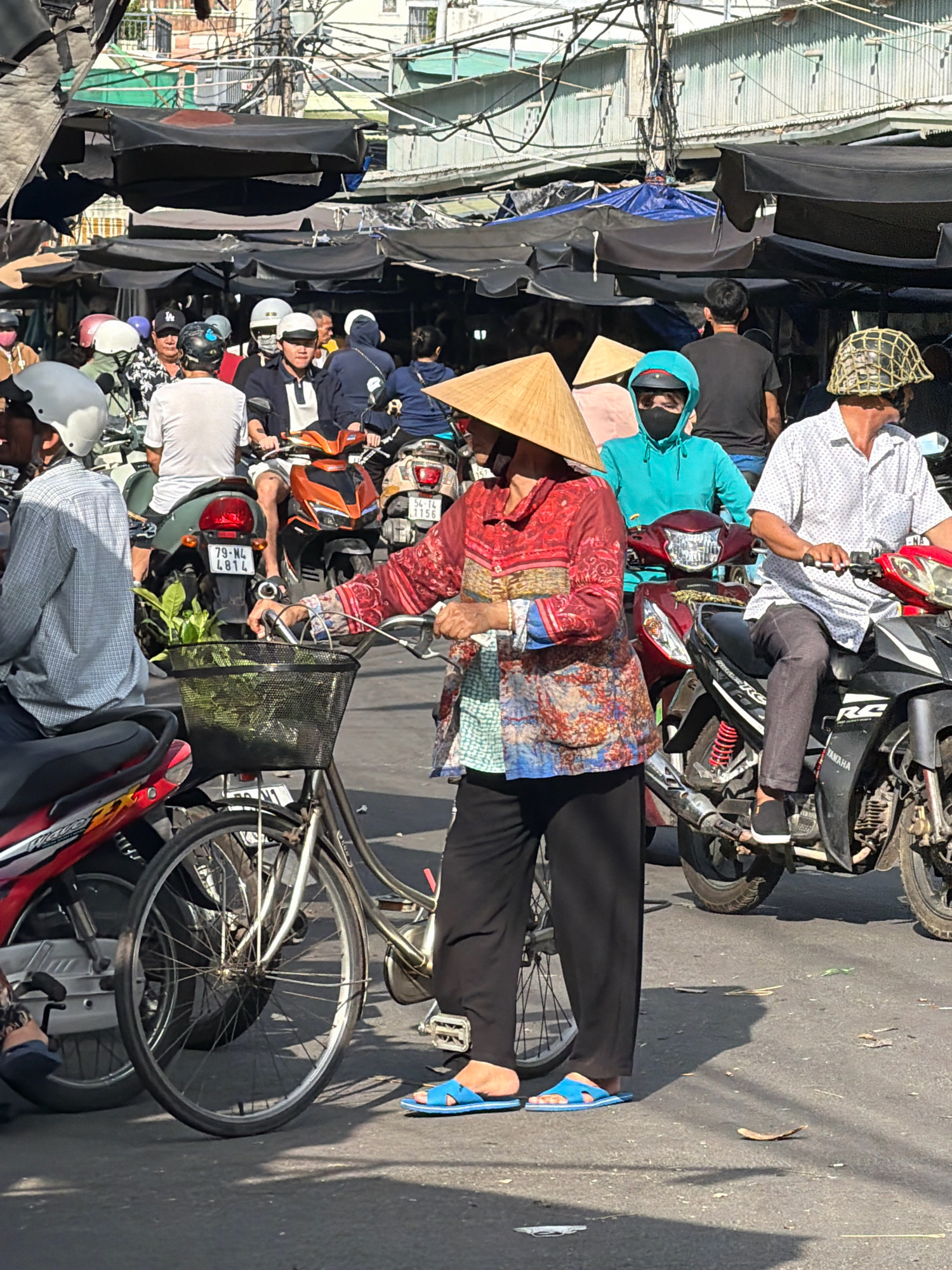The image depicts a busy street market scene in Nha Trang, Vietnam, likely during daylight hours, with overcast skies suggested by the lighting. The foreground features a woman wearing a traditional conical hat and a colorful, patterned long-sleeved shirt, standing next to a bicycle. She is holding the handlebars of the bicycle, which has a black wire basket containing green leafy produce. She is wearing black pants and blue flip-flops. Behind her, several motorbikes are parked or in motion, forming a dense traffic flow.
In the background, a variety of market stalls with dark fabric coverings are visible, casting shadows. People are seen on motorbikes and standing, many wearing helmets and masks, indicating common practice for traffic safety and possibly health precautions. One man on a motorbike wears a distinctive woven helmet. The overall environment suggests an urban setting with crowded conditions, typical of a Vietnamese market. The presence of electric wires overhead further emphasizes the urban infrastructure. The general atmosphere is one of active commerce and daily life.
