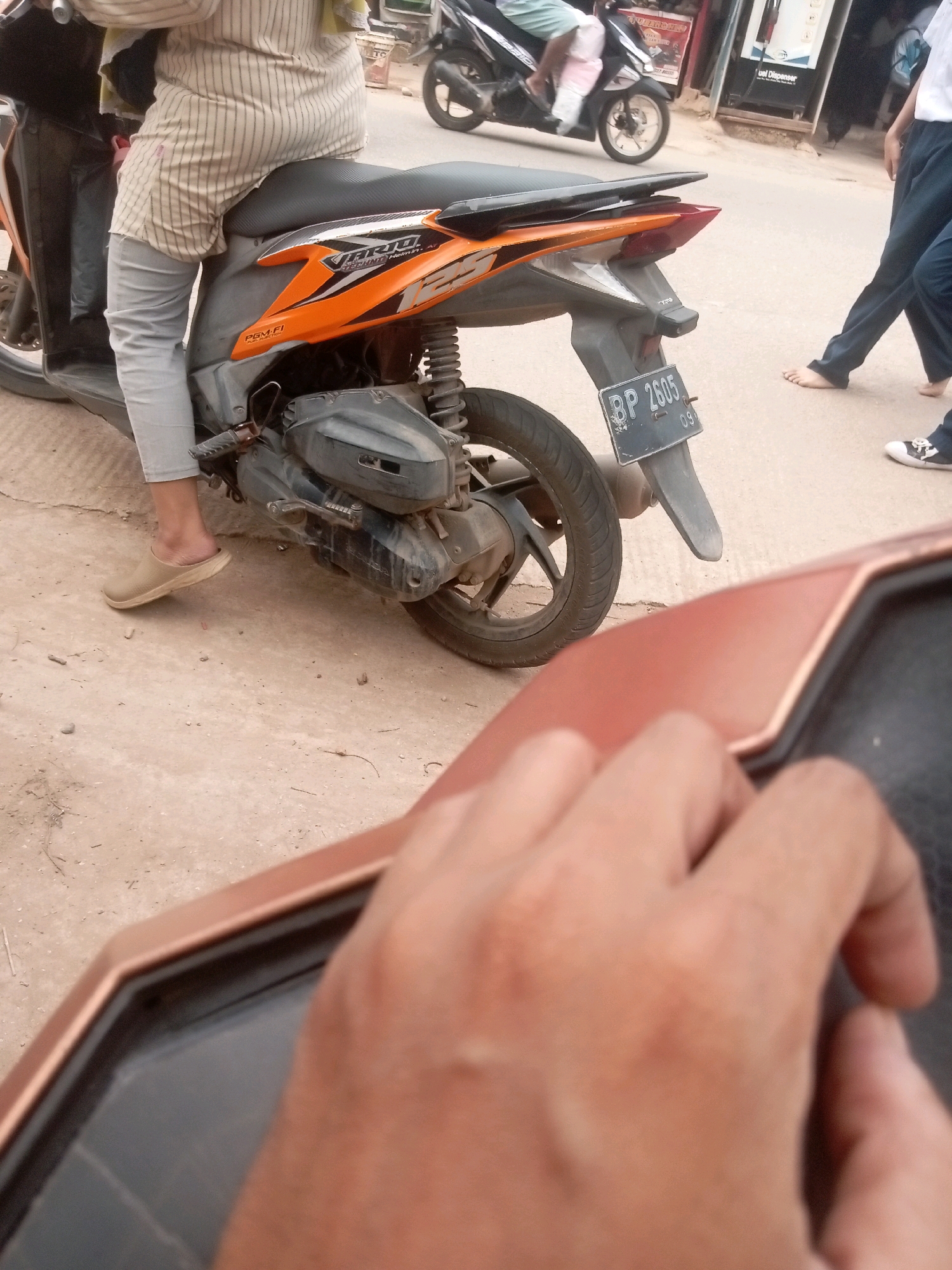 The image shows a person seated on a motorcycle. The person is wearing light-colored pants and a striped shirt. The motorcycle is orange and black. A license plate on the motorcycle reads "BP 2605". In the background, another motorcycle and two people are visible. One person appears to be walking barefoot, while the other is wearing sneakers. The scene takes place on a paved street. This image appears to have been taken in Batam, Indonesia, based on the license plate format and the overall setting.