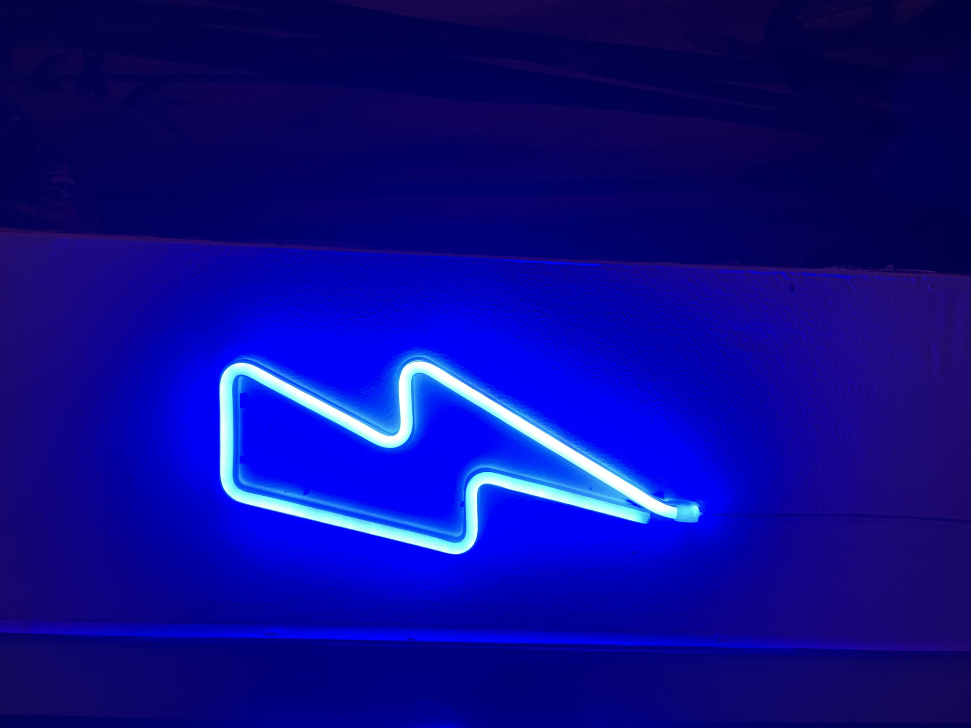 A brightly illuminated blue neon lightning bolt sign is mounted horizontally on a textured, white surface, likely a wall. The surrounding environment is primarily dark blue, with a darker area above the sign suggesting a ceiling or overhang. A thin wire extends from the right end of the sign, indicating its power source. The overall impression is one of artificial lighting in a possibly confined space. Location in Buduk, Indonesia is unknown.