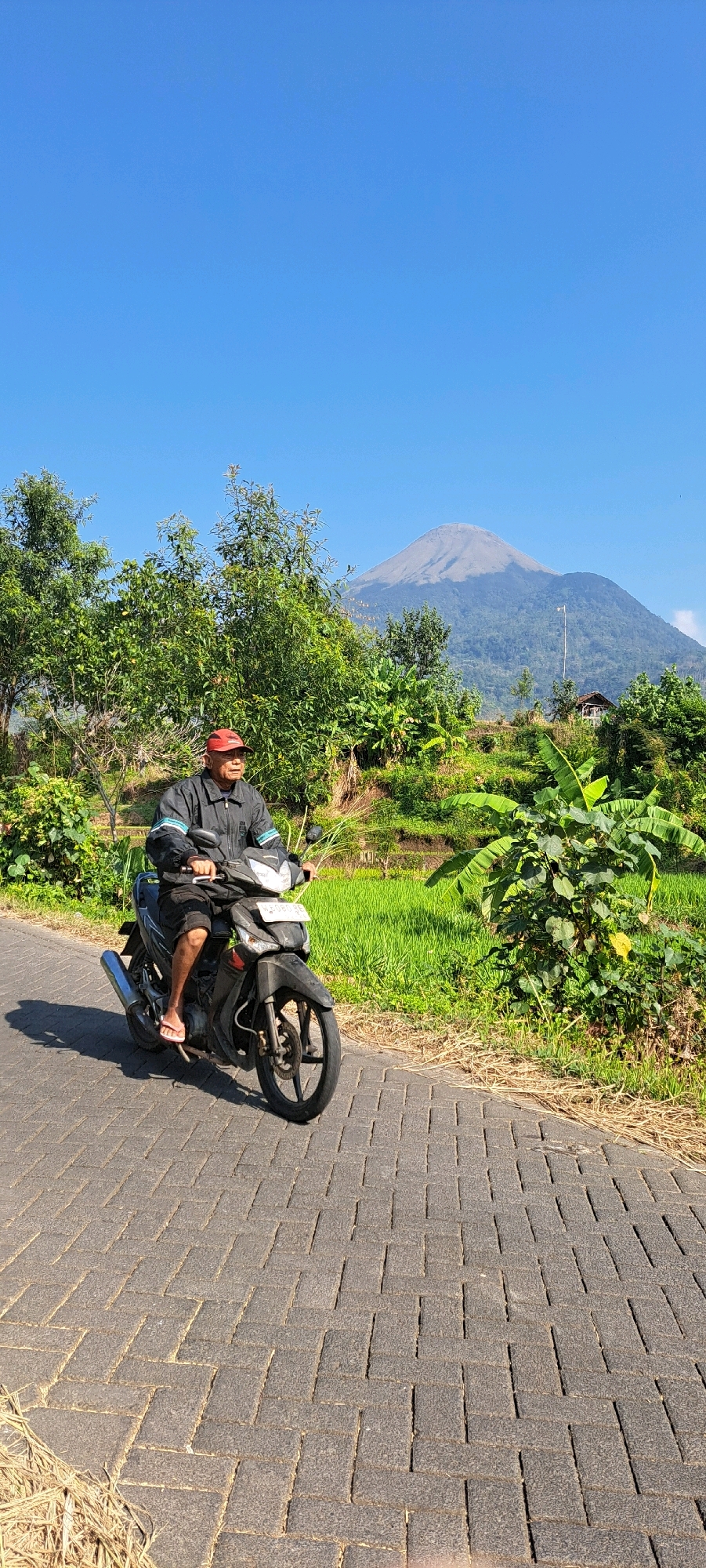 A man wearing a red cap and a dark jacket is riding a black motorcycle on a paved road in Sukoreno, Indonesia. He is wearing shorts and flip-flops. The road is lined with green grass and trees. In the background, there is a mountain under a clear blue sky. The scene appears to be a rural area with rice paddies.