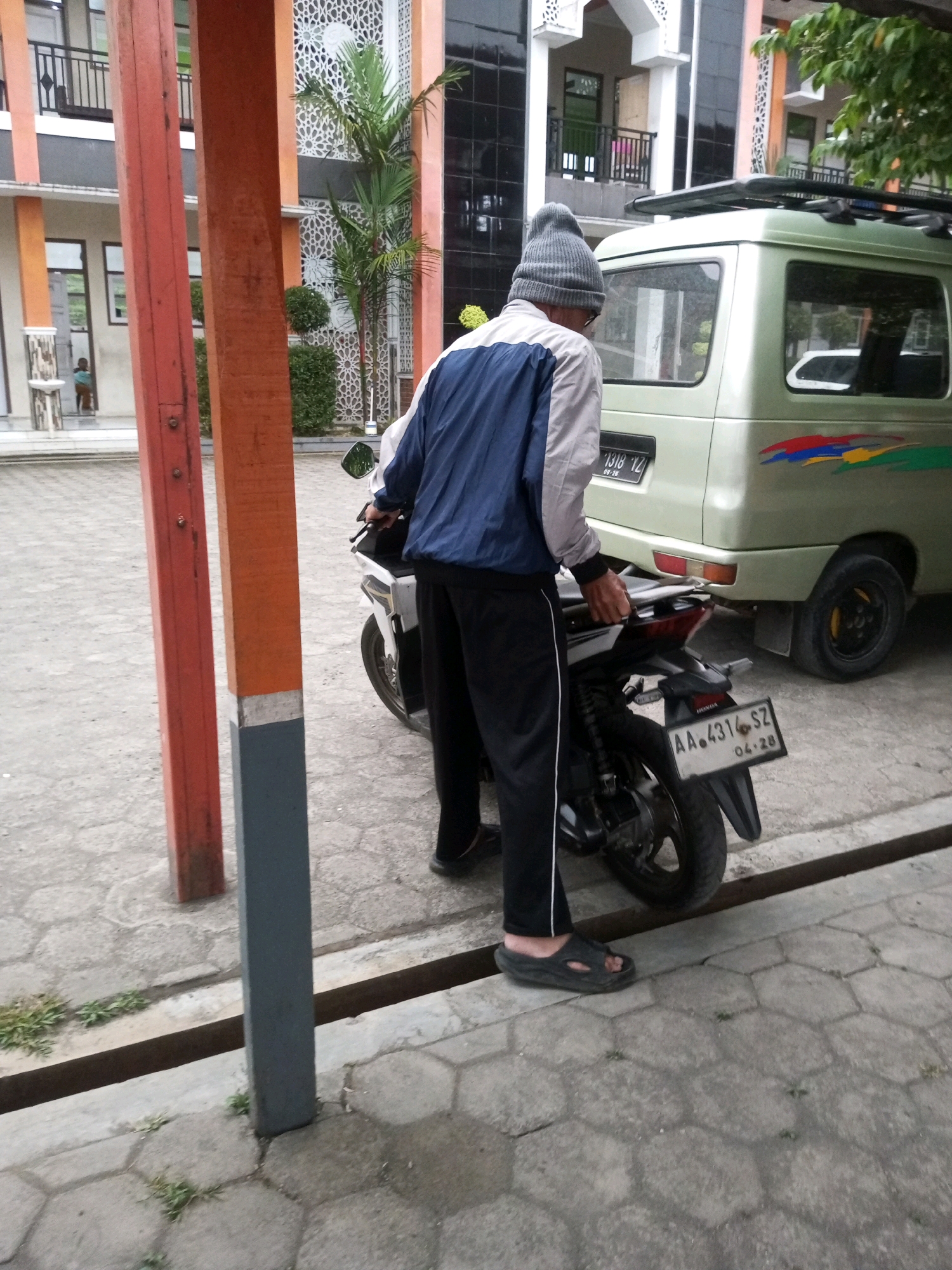 In the city of Kalibeber, Indonesia, a man wearing a grey beanie, a light-colored jacket with dark blue sleeves, and black pants stands next to a white motorcycle. He is holding onto the handlebars of the motorcycle, which has a license plate reading "AA 431 SZ". Behind the motorcycle is a light green van with a license plate "1318 YZ". The van has colorful decals on its side. The scene takes place on a paved ground with hexagonal tiles. In the background, there is a building with orange and white accents, and some greenery. The environment appears to be outdoors, possibly in a residential or public area.