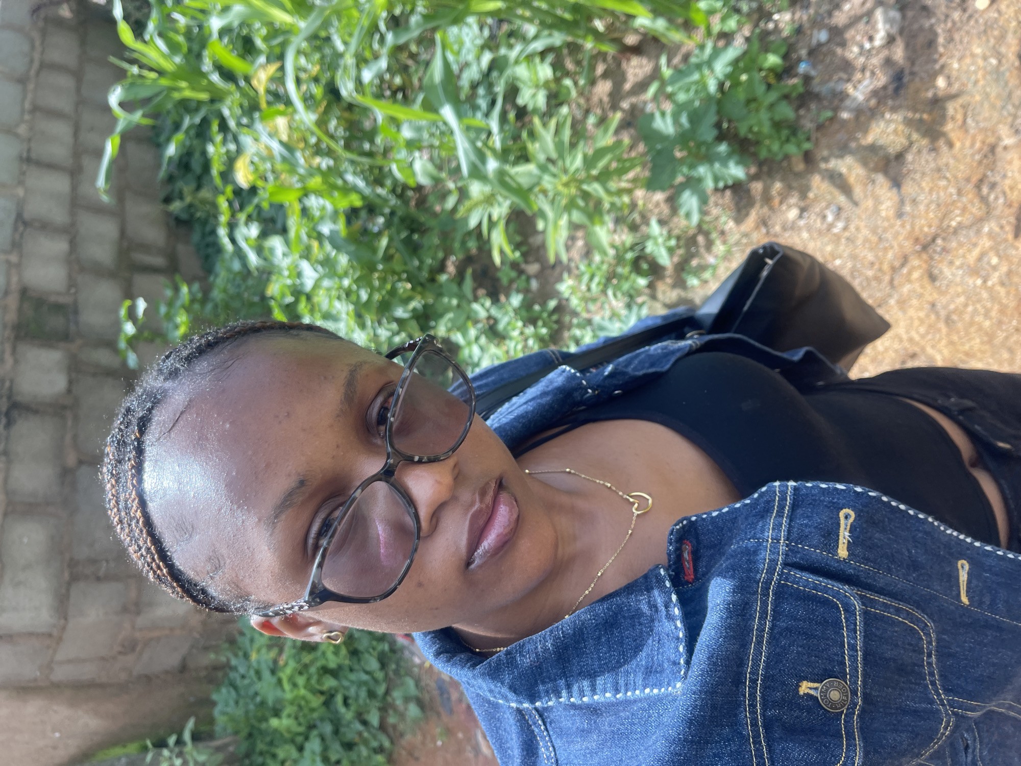 A young Black woman is taking a selfie in an outdoor setting, possibly in Rafin Par, Nigeria, as indicated by the presence of lush greenery and what appears to be a brick wall in the background, common in many Nigerian communities. She is wearing a blue denim jacket over a black top and a delicate gold necklace with a heart pendant. She also has on dark-rimmed glasses and small hoop earrings. Her hair is styled in thin braids that are pulled back from her face. The ground is a mix of dirt and scattered pebbles. The overall impression is casual and natural, with the subject seemingly relaxed and confident.