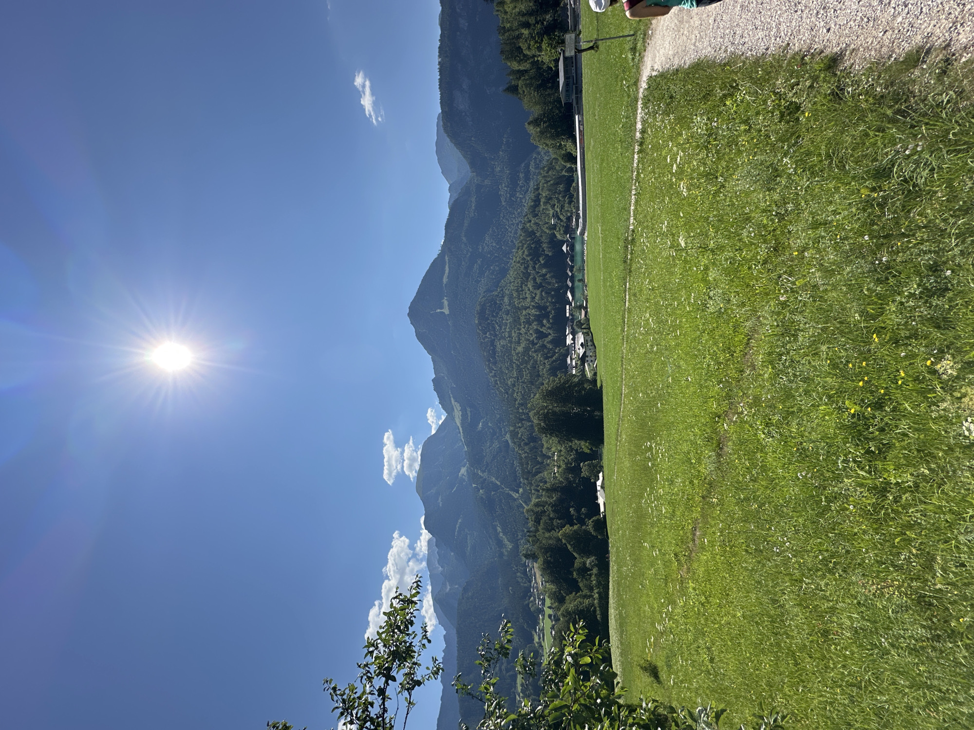 This is a wide-angle, eye-level, outdoor photograph taken on a sunny day in Schönau am Königssee, Germany. The scene depicts a vibrant green meadow dotted with small white and yellow wildflowers, leading to a light-colored gravel path on the right. In the background, lush green trees cover rolling hills, which transition into majestic, imposing mountains with visible ski slopes. A small village with buildings and a body of turquoise water are nestled at the base of the mountains. The sky is a clear, bright blue, dominated by the sun, which shines intensely and creates a lens flare effect. A person wearing a white hat and a teal top is visible on the right side of the path, walking away from the camera. The image captures a scenic view of the Bavarian Alps, a popular tourist destination known for its natural beauty and outdoor activities.