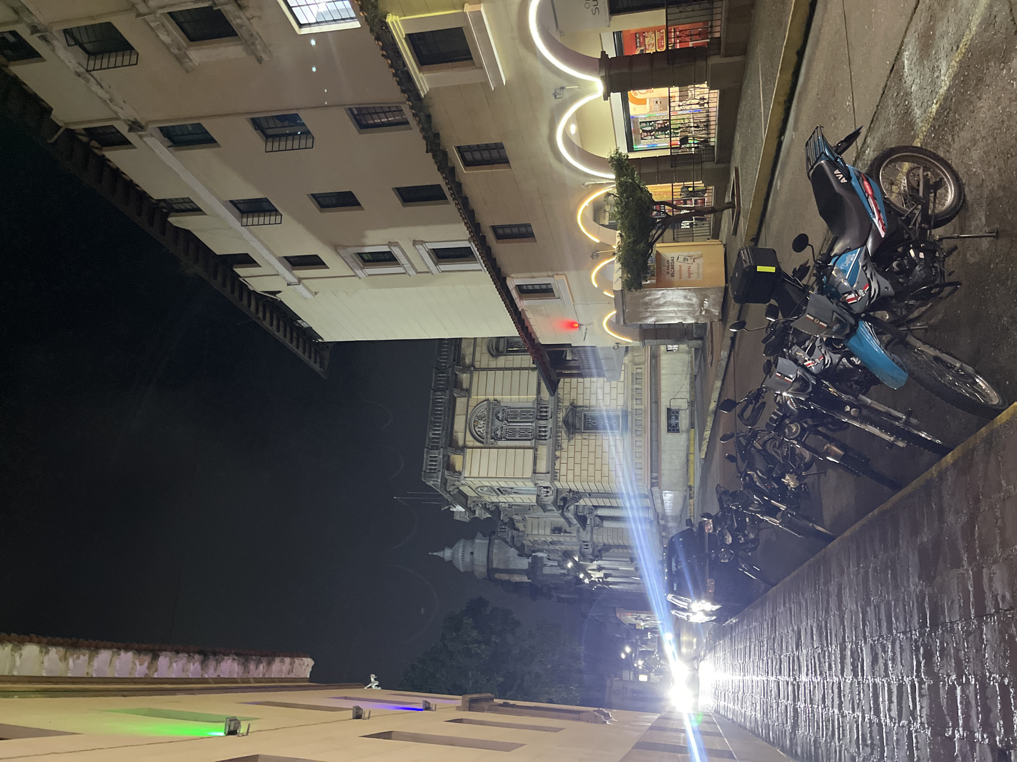A nighttime, low-angle shot captures a street scene in Mérida, Venezuela. The street is paved with brickwork, wet from recent rain, reflecting the ambient light. A row of parked motorcycles lines the right side of the road, their forms illuminated by the harsh glare of headlights from an unseen vehicle approaching from the left. The headlights create long, streaking lens flares across the image.
On the left, the side of a light-colored building is visible, featuring a green light fixture near the top. Beyond the parked motorcycles, the street curves slightly, revealing more of the urban environment. Across the street, a building with arched walkways and illuminated by warm-toned lights stands prominently. Shops or businesses appear to be housed within these arches. Further down the street, a grand, ornate building with classical architectural elements is partially visible, its facade illuminated by streetlights. A domed tower is discernible above this building, suggesting a significant landmark. The sky is dark and overcast, typical of a nighttime city scene. No people are clearly visible in the frame, and the focus is on the static elements of the street and architecture.
