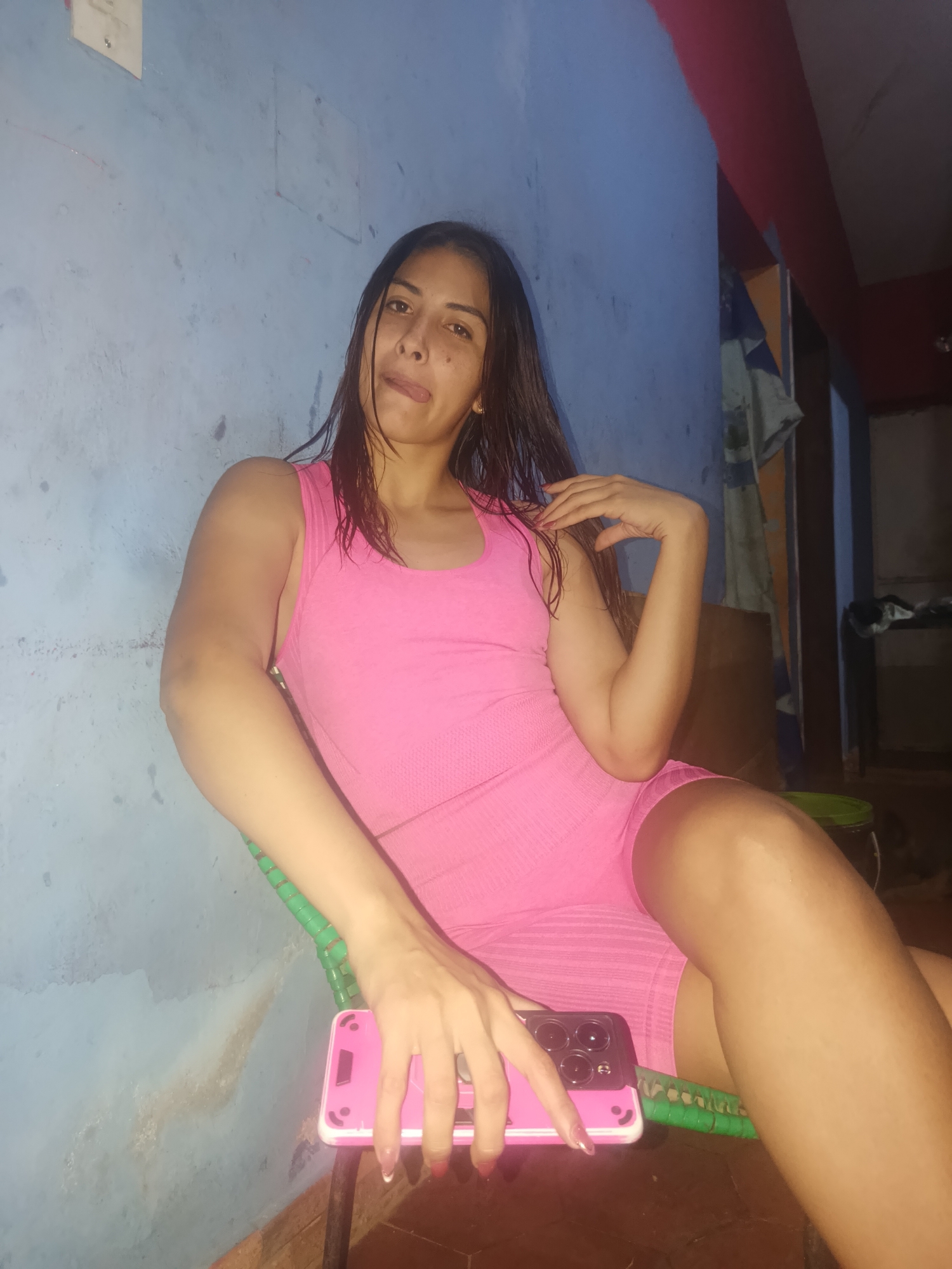 A young woman with long, dark, slightly wet hair is seated, posing for the camera indoors in Juan Griego, Venezuela. She wears a form-fitting, bright pink one-piece outfit consisting of a tank top and shorts. Her left hand rests on her lap, holding a pink smartphone with multiple camera lenses. Her right hand is relaxed by her side, touching her shoulder. She is looking directly at the camera with a playful expression, her tongue slightly extended.
The woman is sitting in a green chair, possibly with a woven or mesh design. The background features light blue walls that show signs of wear, including discoloration and possible dampness or peeling paint in various areas. A red beam or trim is visible towards the top right, and some draped fabric or clothing can be seen in the dimly lit background on the right side. The overall lighting suggests an indoor setting, possibly during evening or night. No visible text is present in the image.