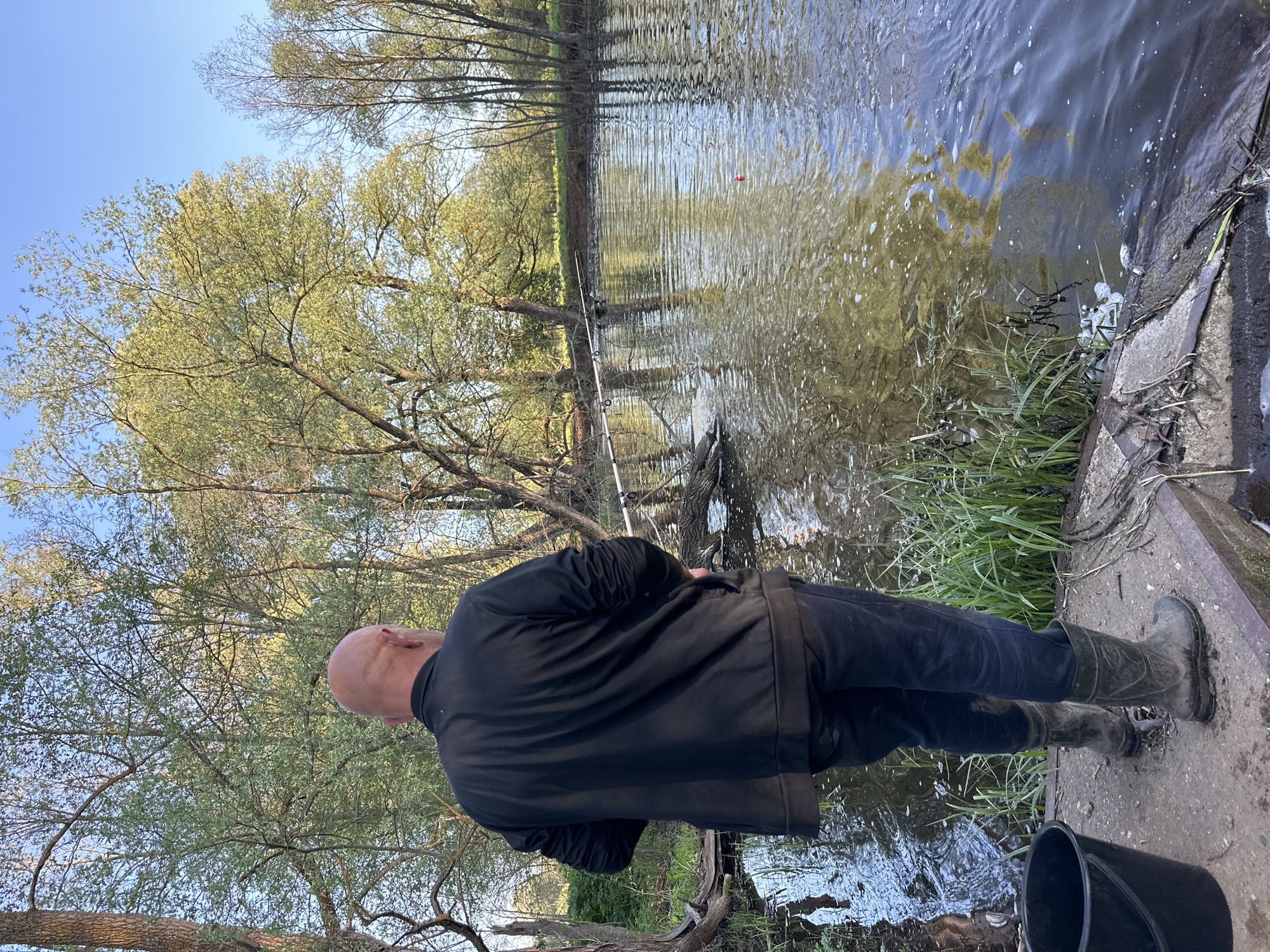 A man stands on a concrete bank next to a body of water. He faces the water and appears to be fishing, though the fishing rod is somewhat obscured by trees in the background. The man is bald, wearing a black jacket, dark pants, and rubber boots. A black bucket sits to his right on the concrete. The water reflects the trees lining the opposite bank. The scene is in Альняш, Russia.
