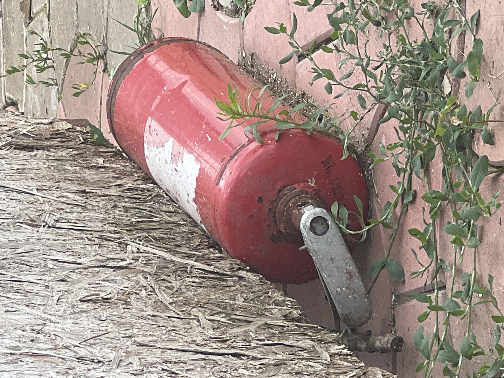 A red fire extinguisher is lying on its side on a brick patio, partially obscured by a large, rough-textured object that appears to be a bale of straw or woodchips. The fire extinguisher is cylindrical with a white stripe on its side and a metal handle mechanism at one end. Small green plants are growing through the cracks in the brickwork around the objects. The scene is outdoors, with natural light casting shadows. The location is identified as Sorochinsk, Russia.