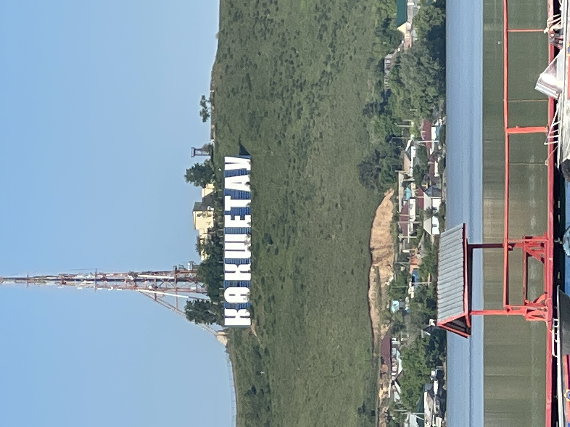 The image shows a view of Kokshetau, Kazakhstan, with the city's name spelled out in large, white Cyrillic letters on a green hillside. A tall, metal communication tower stands to the left of the sign. Below the hillside is a residential area with various houses and trees. A body of water is visible in the foreground and to the right of the hillside, and part of a structure with red supports is on the right edge of the image. The sky is clear and blue.