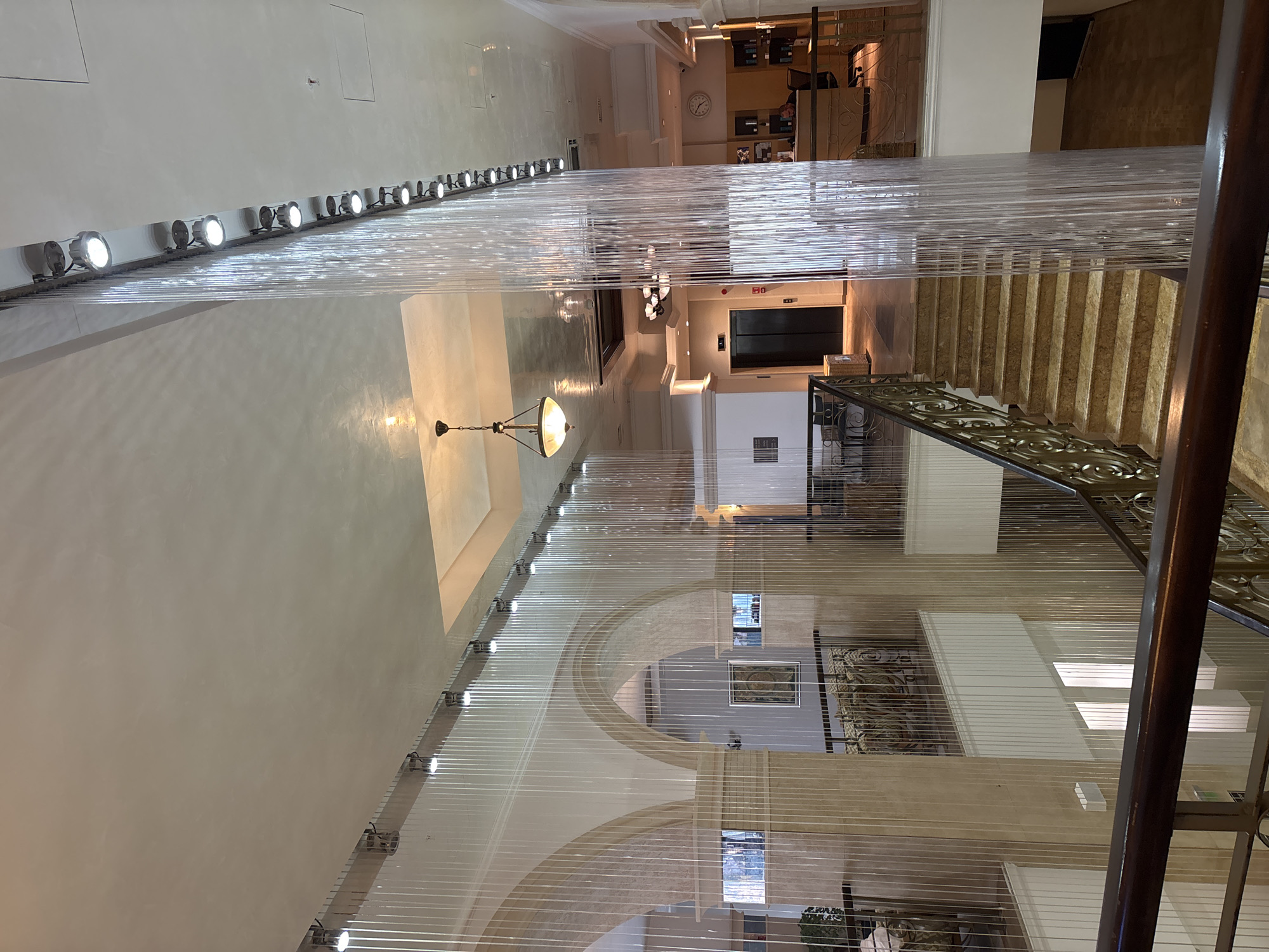 The image depicts the interior of a multi-level building, likely a hotel or a commercial establishment, with a grand staircase as a central feature. The architecture incorporates arched openings and marble accents. A prominent design element is a vertical waterfall of clear, transparent strands illuminated by a row of spotlights along the ceiling, creating a shimmering effect.
The ground floor area is visible, with an elevator, a reception desk manned by a person, and display shelves. On the upper levels, balconies overlook the central space, furnished with chairs and tables. The overall aesthetic suggests a modern and somewhat luxurious environment. The image does not provide any visual cues to confirm its location in the city of Razliv, Bulgaria, or any other specific geographical area.