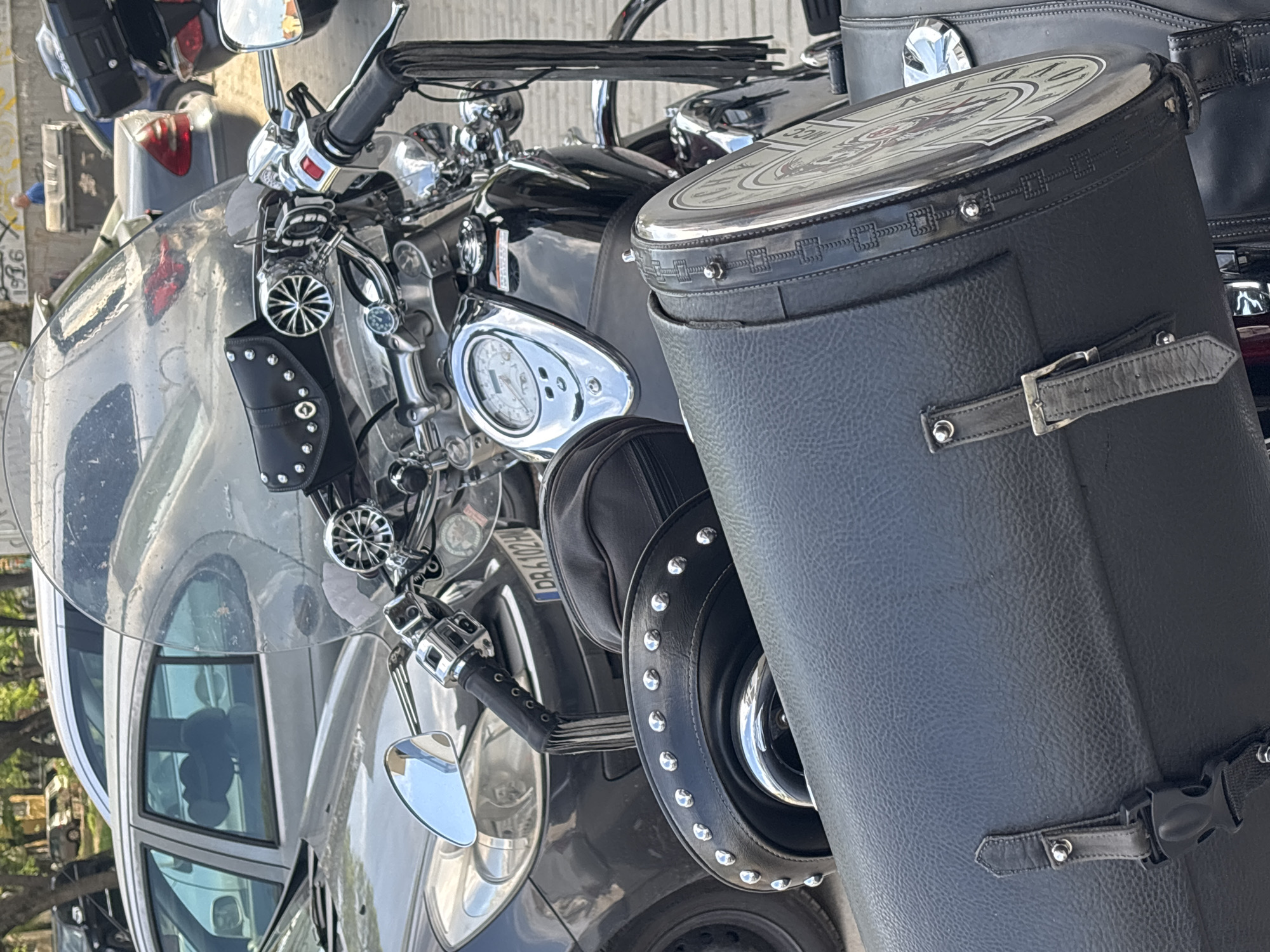 A black cruiser motorcycle is prominently featured, parked outdoors during the daytime. The motorcycle is adorned with extensive chrome accents on its handlebars, controls, and mirrors. A large, clear windshield is visible at the front, reflecting parts of the surroundings, including a silver car parked alongside.
Notable features include black leather accessories such as a small studded pouch on the handlebars and a large, cylindrical black leather storage bag, possibly a saddlebag, attached to the side. This cylindrical bag has a decorative metallic lid featuring an ornate crest design that appears to include a lion-like figure, and some indistinct text or numbers, possibly "MCC" and a year. The bag is secured with leather straps, buckles, and a plastic clasp. The front wheel is partially visible, covered in black leather with a studded rim.
A Bulgarian license plate, "DR 102BH", is visible, confirming the location as Plovdiv, Bulgaria. The motorcycle's dashboard includes a circular speedometer with chrome trim. Reflections in the car's windows show hints of the environment and a vague reflection of a person. The bright sunlight indicates clear weather. The motorcycle is stationary, with no specific activity taking place.