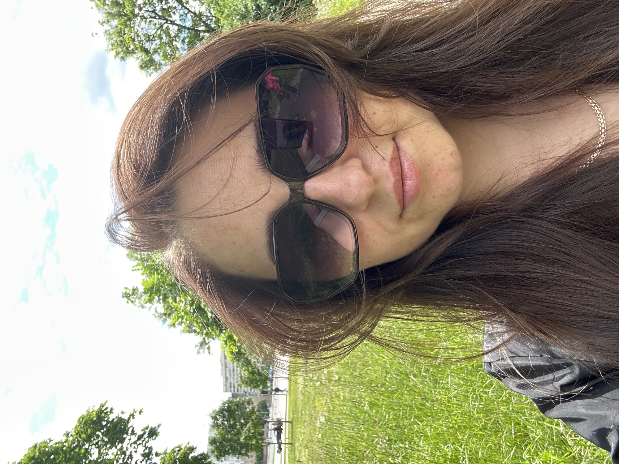 A close-up, head-and-shoulders selfie taken outdoors on a sunny day. The subject, a woman with long, brown hair and wearing large, dark sunglasses, is positioned slightly off-center to the right. Her face is mostly visible, with her lips slightly parted and a gentle smile. Her skin shows a few freckles. She is wearing a dark garment, possibly a black jacket or shirt, and a gold-colored necklace. Her hair is wind-blown, with strands falling across her face and shoulders.
The background features a lush green grassy field with some sparse wildflowers. Beyond the field, trees with vibrant green leaves are visible, and further in the distance, there are buildings with a light-colored facade, suggesting an urban or suburban setting. The sky above is bright blue with scattered white clouds. Reflections in the sunglasses show a person in a pink outfit holding a phone, likely the photographer. The overall impression is a casual, outdoor portrait.