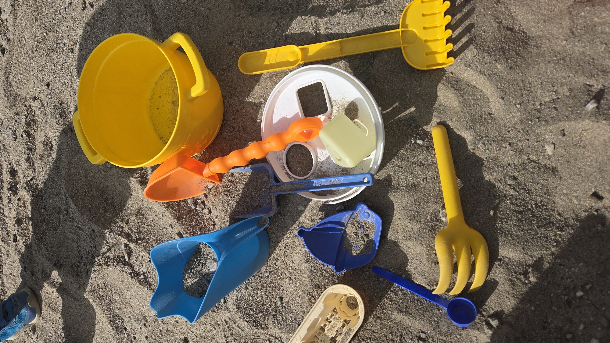 In Tynset, Norway, a collection of colorful plastic sand toys lies scattered on a sandy surface. The toys include a large yellow bucket with two handles, an orange scoop, a blue shovel with a broad, angled head, a blue bucket mold shaped like a hat, a yellow rake, a yellow small shovel and a blue small spoon. A white circular lid with square and circular openings sits in the center of the arrangement, with a small, light yellow rectangular object resting on it. The sand is a light brown color with small pebbles mixed in. In the upper left corner, the lower portion of a blue and black sneaker is visible, suggesting a person was present at the scene. The scene is illuminated by sunlight, casting shadows from the objects onto the sand.