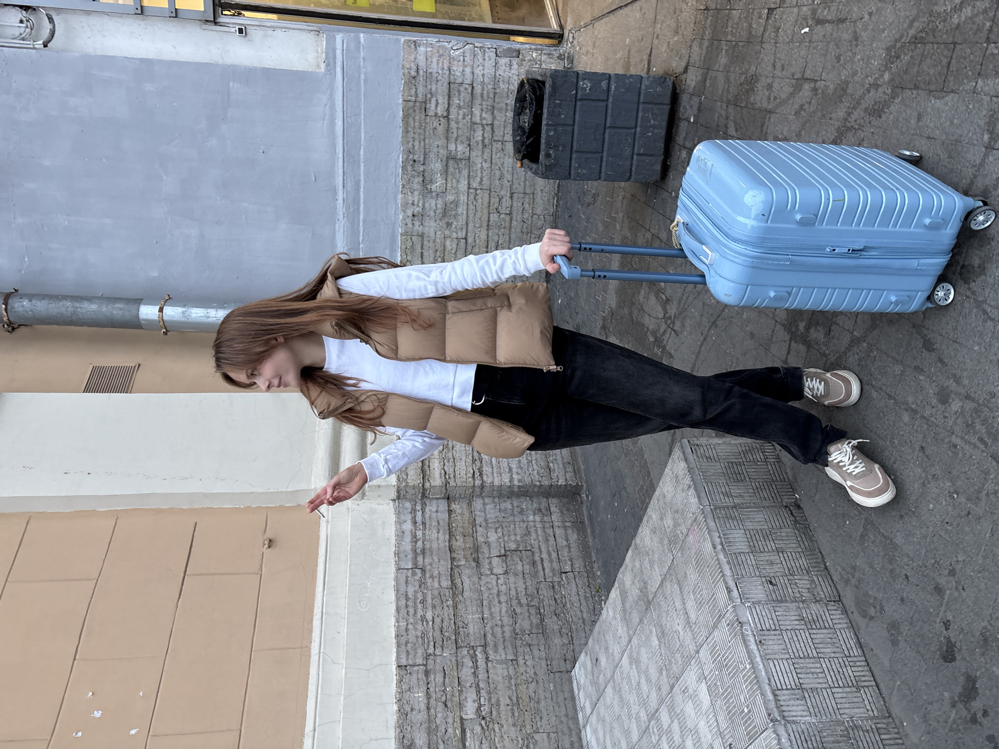 A young woman is standing on a sidewalk in Saint Petersburg, Russia, holding a lit cigarette in her right hand and pulling a light blue suitcase with her left hand. She wears a white, long-sleeved shirt, a tan puffer vest, black jeans, and taupe-colored sneakers. The background includes a yellow and white building, a gray building, a gray brick wall, a black trash bin, and gray concrete tiles. The scene appears to be outdoors, possibly near a commercial area.