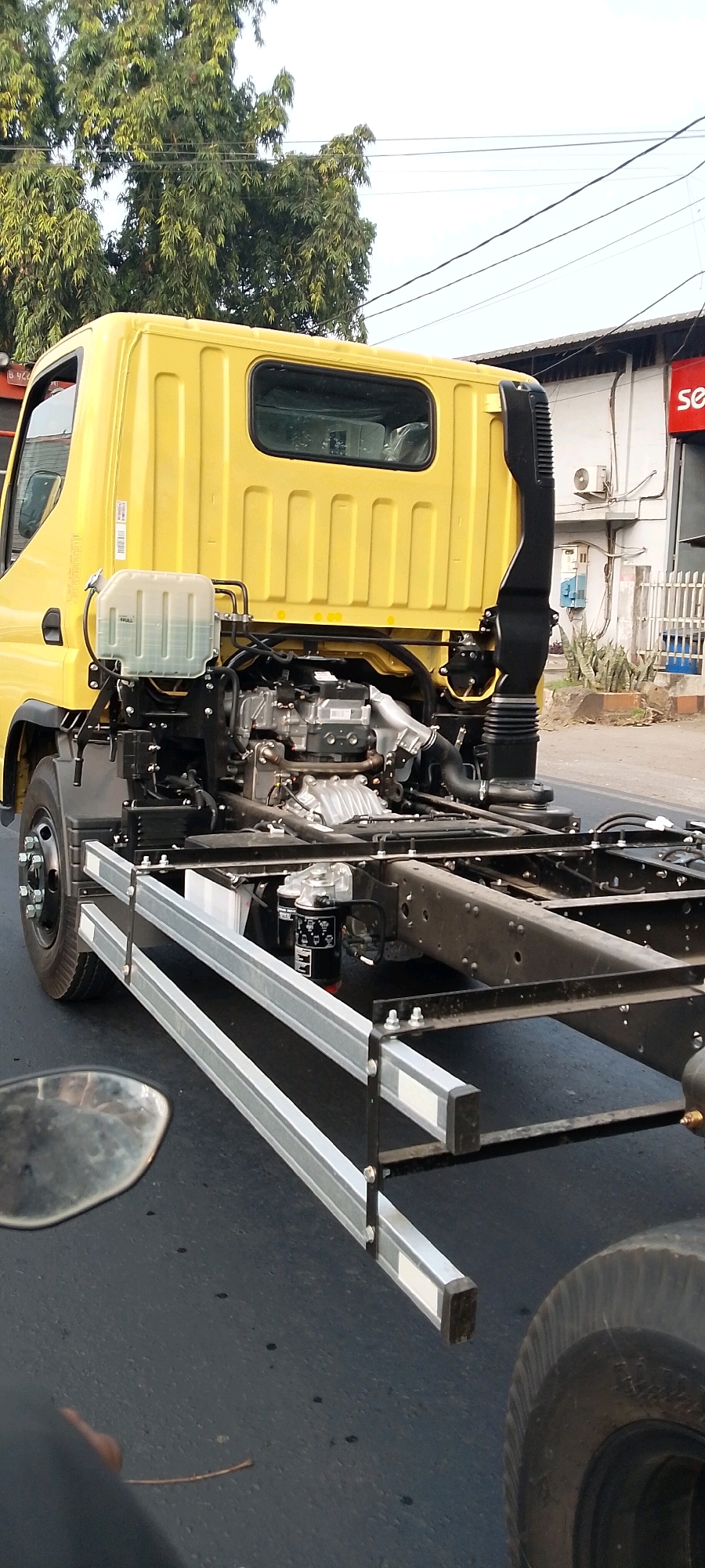 The image shows the rear chassis of a yellow Fuso truck, likely a new model under assembly or transport. The truck is on a street in Bekasi, Indonesia. The engine and exhaust system are visible. The truck is stationary, and there are no people in the immediate vicinity. The background includes trees, power lines, and buildings, suggesting an urban or semi-urban environment. The road surface is asphalt, and there is a side mirror and a tire from another vehicle in the foreground, indicating the photo was taken from another vehicle.