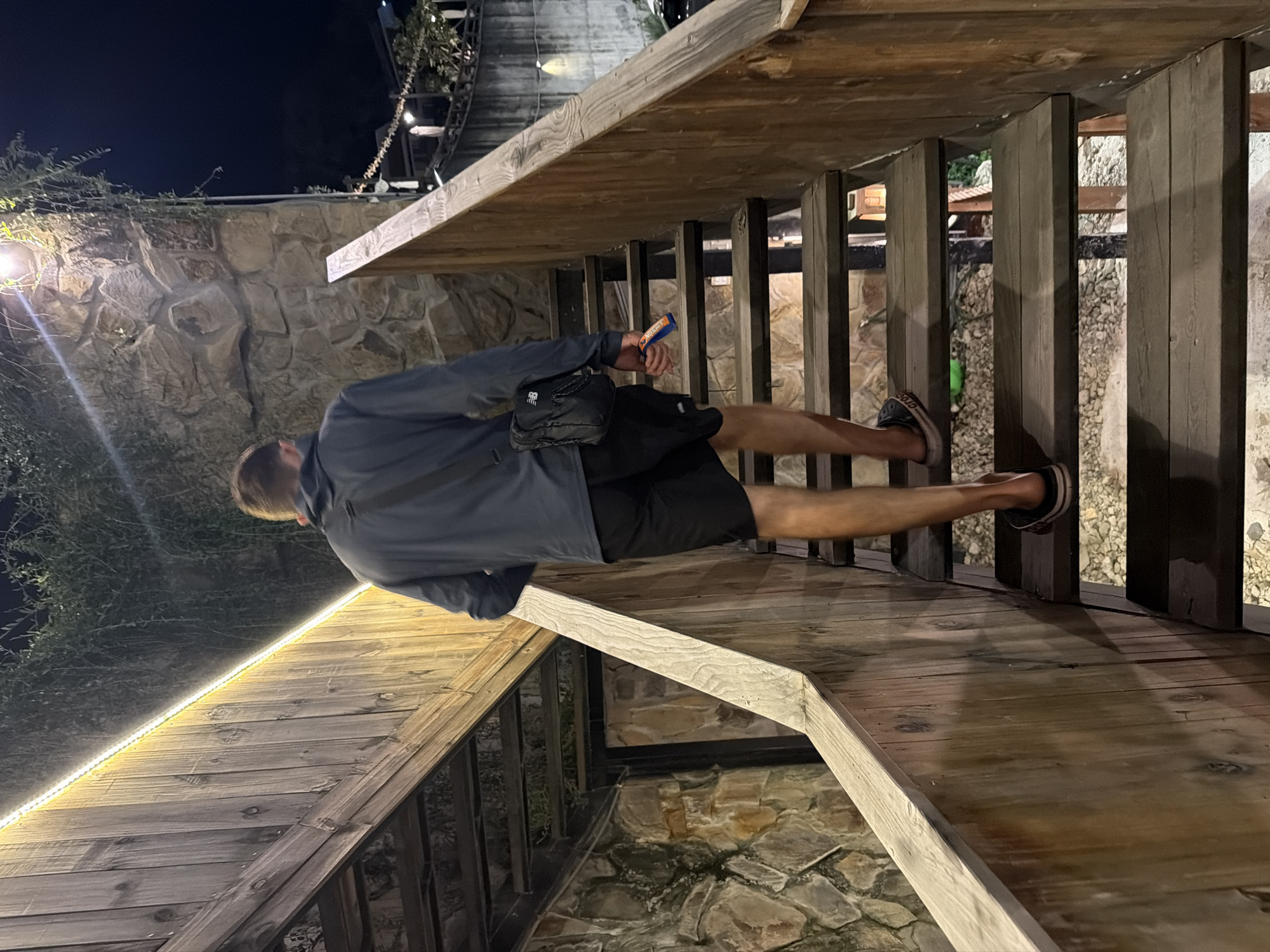 The image depicts a male individual ascending a wooden outdoor staircase at night in Nam Nha Trang Ward, Vietnam. The man, viewed from behind, wears a dark long-sleeved shirt, dark shorts, and dark sandals. He carries a black New Balance cross-body bag on his left hip and holds a blue and orange object in his right hand. The staircase is constructed from dark wooden steps and light-toned wooden railings on both sides. A linear warm LED light strip illuminates the underside of an overhead wooden structure running parallel to the stairs. To the left, a textured stone wall with trailing green vegetation is visible, featuring an additional mounted light fixture. Rocky ground is discernible through the open risers of the wooden steps. The distant background is dark, indicating nighttime, with faint artificial lighting.