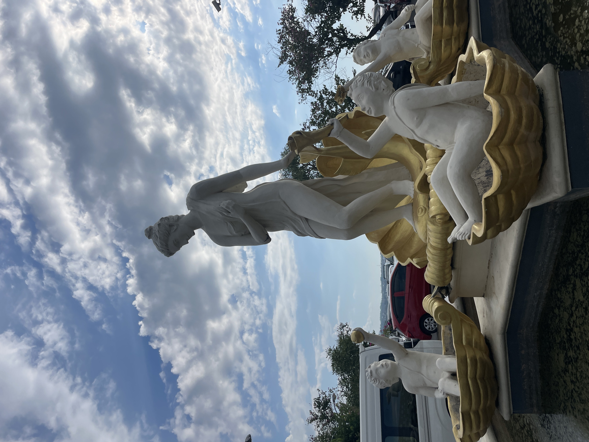 A white sculpture of a female figure standing on a large golden shell is the central subject of this outdoor scene. She is draped in flowing fabric and appears to be looking downwards. Around the base of the central figure, four cherubic statues are seated in smaller golden shell-like vessels. One cherub on the left holds a golden orb in its raised hand. Another cherub in the foreground holds a golden object to its mouth. The background includes trees, a red car, a white van, and other parked vehicles, suggesting a public or accessible area. The sky above is a bright blue with scattered white clouds. This fountain sculpture is located at the Boulevard Area in Manado, Indonesia.