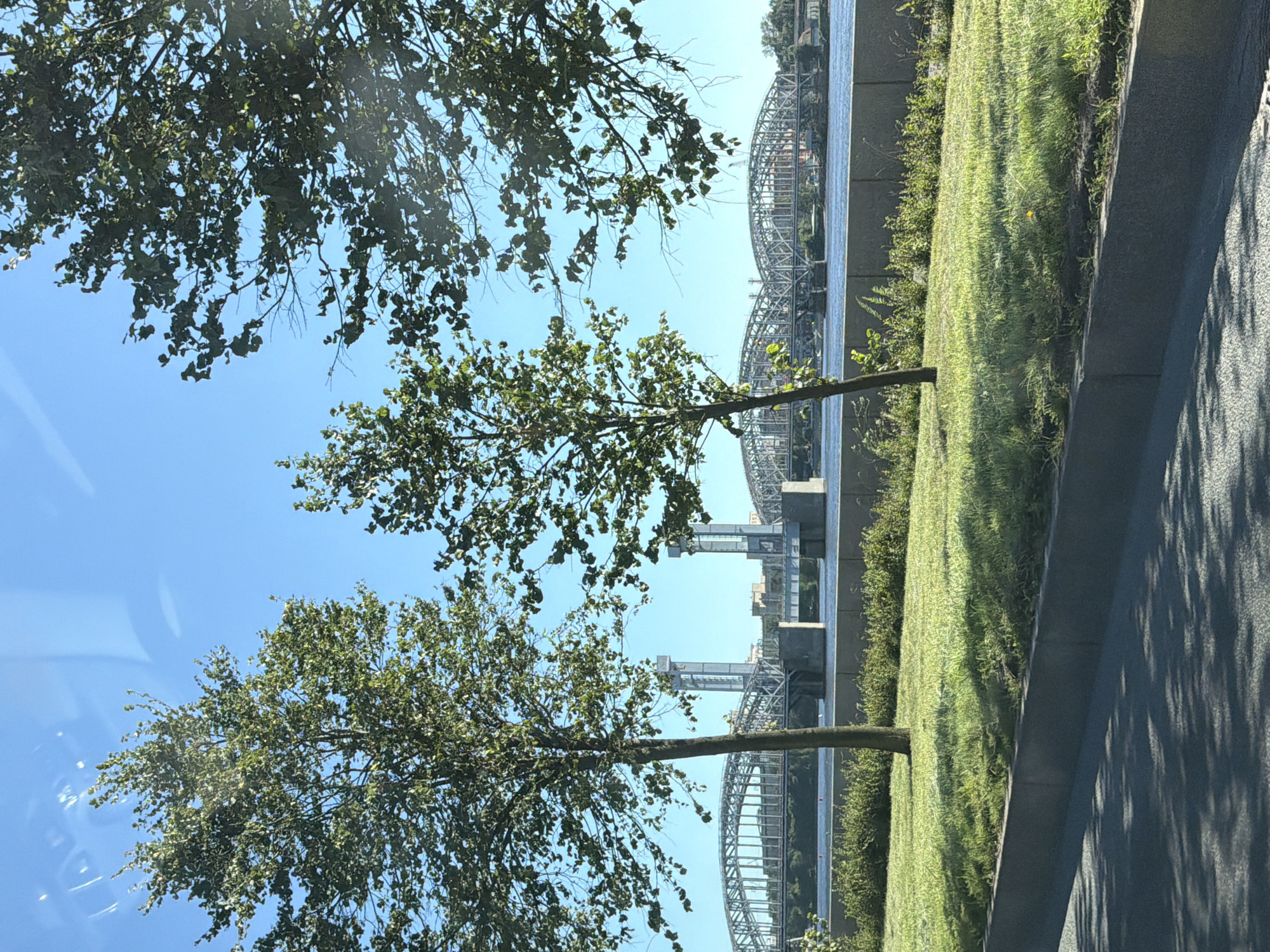 The image shows a view of the Bolsheokhtinsky Bridge in Saint Petersburg, Russia, on a clear day. The bridge, a prominent landmark, is a metal arch bridge spanning the Neva River. Two tall, industrial-style towers flank the central section of the bridge, which appears to be a drawbridge mechanism. The water of the Neva River is visible in the foreground, with a grassy embankment and some trees in front of the bridge. The embankment is bordered by a concrete structure and a paved surface, possibly a road or sidewalk, which is shaded by the foliage of trees. The sky is a vibrant blue. No people are visible in the image. The scene is calm and depicts an urban landscape with significant architectural and natural elements.