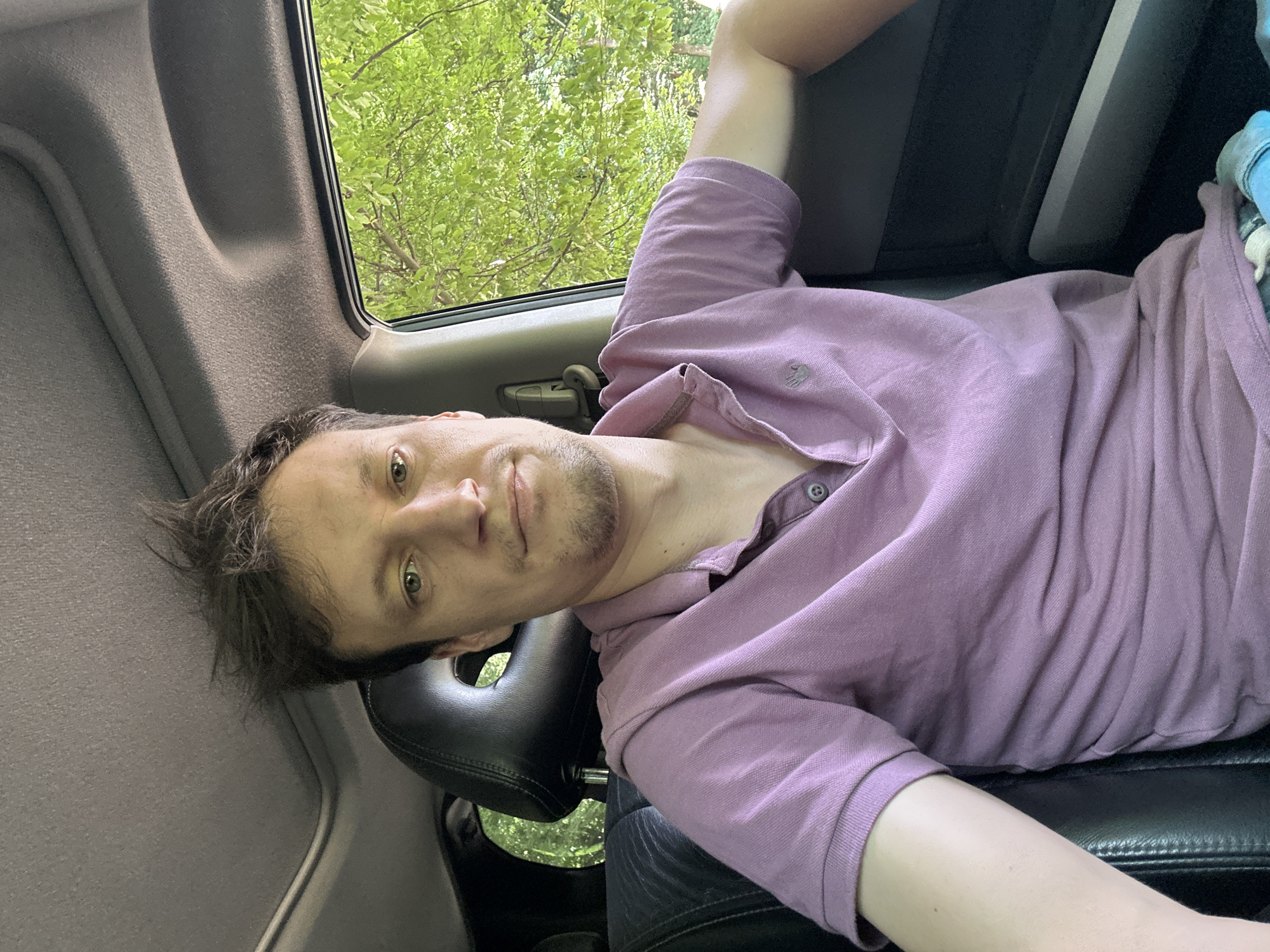 A man with tousled brown hair is seated in the passenger seat of a car, looking directly at the camera. He is wearing a light purple polo shirt. The car's interior is visible, including a black headrest and a grey headliner. Outside the car window, lush green trees can be seen, suggesting an outdoor setting likely within the city of Shatava, Ukraine. The man's facial expression is neutral.