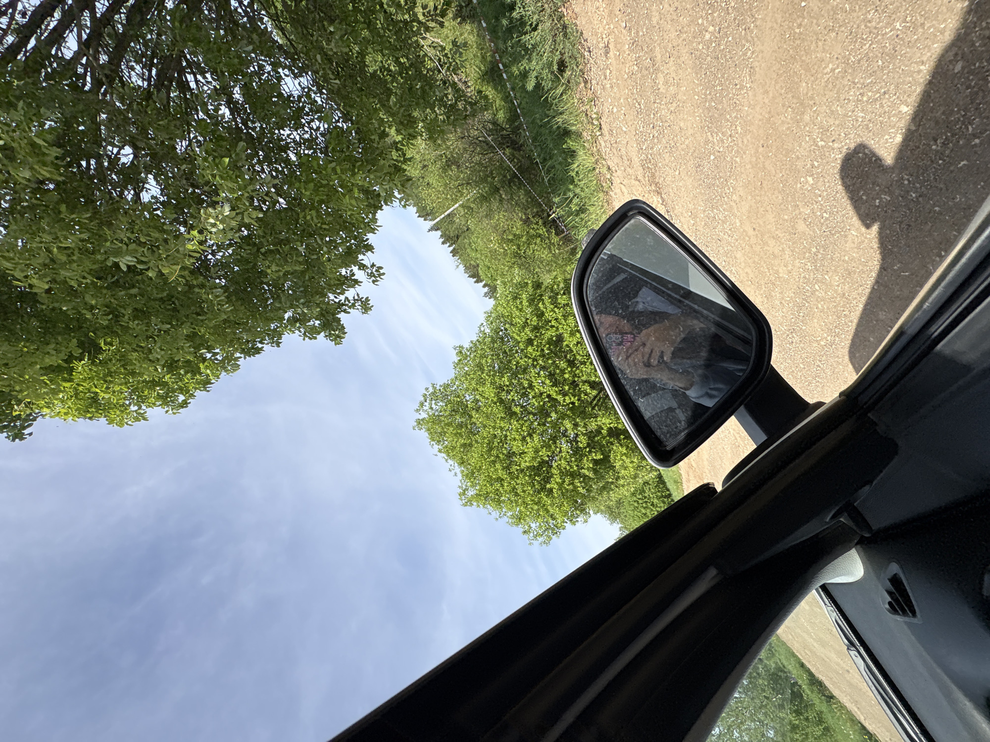 The image appears to be taken from inside a car, looking out and upward at an angle. The visible environment is dominated by trees with lush green foliage and a bright sky with scattered clouds. The car's side mirror is prominent, reflecting what appears to be a person with pink hair inside the car. A dirt road is visible along the right side of the frame, with the shadow of a hand also being seen on the dirt. The interior of the car, including the window frame and a portion of the dashboard, is black. There is no clear evidence to determine the specific location in the city of Sibir', Russia.