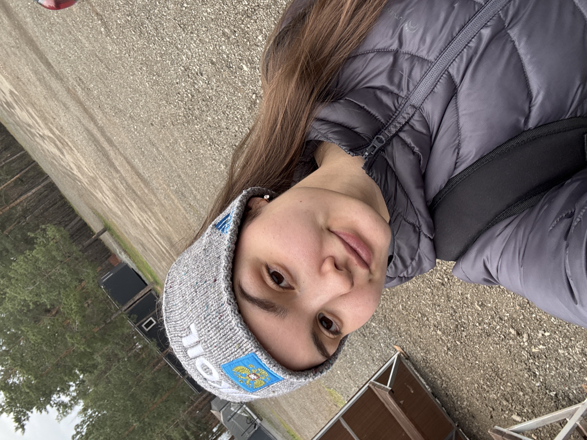 A young woman, likely in her late teens or early twenties, takes a close-up selfie in an outdoor setting in Orenburg, Russia. She is oriented sideways in the image, looking directly at the camera with a subtle, content smile. Her long, brown hair falls over her shoulder, and she wears a small, sparkling earring.
She is dressed for cool weather in a grey quilted puffer jacket and a grey knit beanie. A black strap, likely from a backpack or bag, is visible across her chest. A prominent feature of her beanie is a blue and yellow patch depicting a double-headed eagle with a shield on its chest, characteristic of Russian heraldry, reinforcing the specified location context. Faint white letters are partially visible on the beanie below the patch.
The background shows a gravel or stone-covered ground and several pine trees, suggesting a natural or possibly an event area. Dark-colored structures, resembling containers or small buildings, and lighter wooden or metal frameworks are seen in the mid-ground. The lighting is natural but appears overcast, indicating daytime.