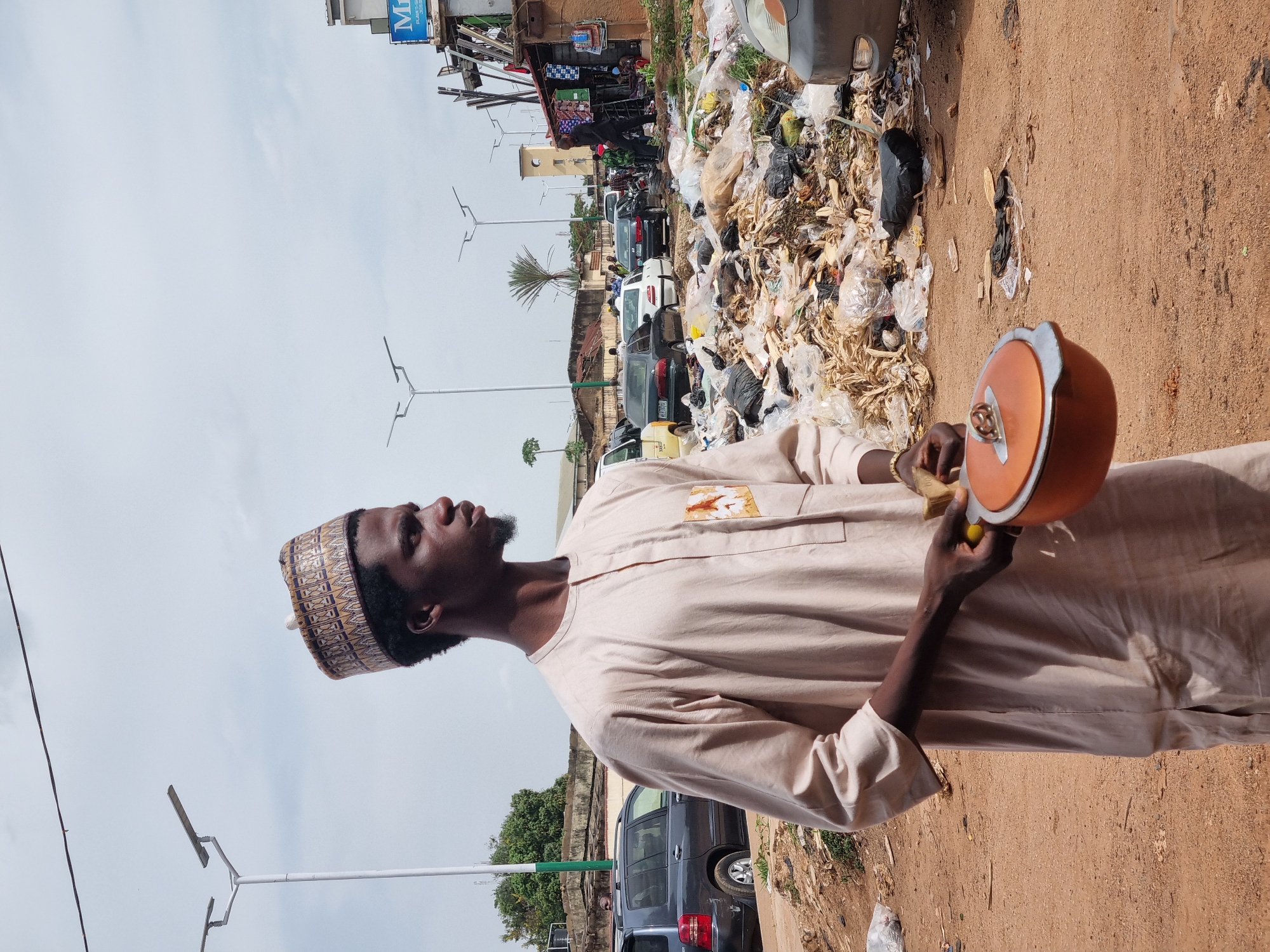 A man wearing a traditional patterned hat and a beige tunic stands on a dirt-covered roadside in Jos, Nigeria, holding an orange pot. Behind him is a large pile of garbage next to a road with parked cars and various shops. Overhead are light poles and power lines.