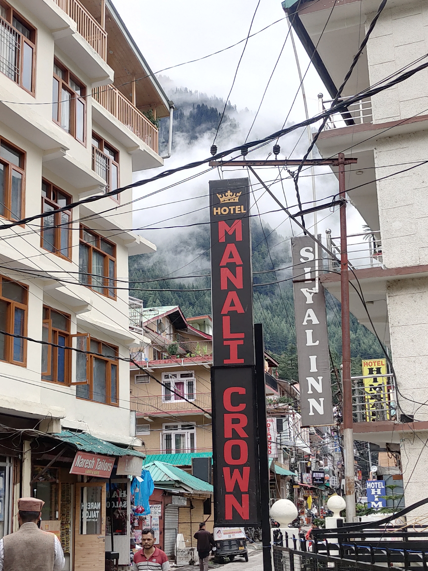 The image shows a street scene in Manali, India, with multiple buildings and signs. A tall, black sign with red lettering that says "MANALI CROWN HOTEL" is prominent in the foreground. To the right of this sign is a gray sign with white lettering that reads "SIYALIN". Further to the right, a yellow sign with black lettering says "HOTEL H M". A white building with multiple windows and balconies is visible on the left. In the background, misty mountains covered in trees are seen. Several electrical wires are strung across the scene. A man wearing a traditional cap and vest is standing on the left, facing away from the camera. Another man in a striped shirt is standing in the middle of the frame. A small shop with a green roof and a sign reading "Naresh Tailors LADIES & GENTS" is also visible. The overall impression is a bustling street in a mountainous town.