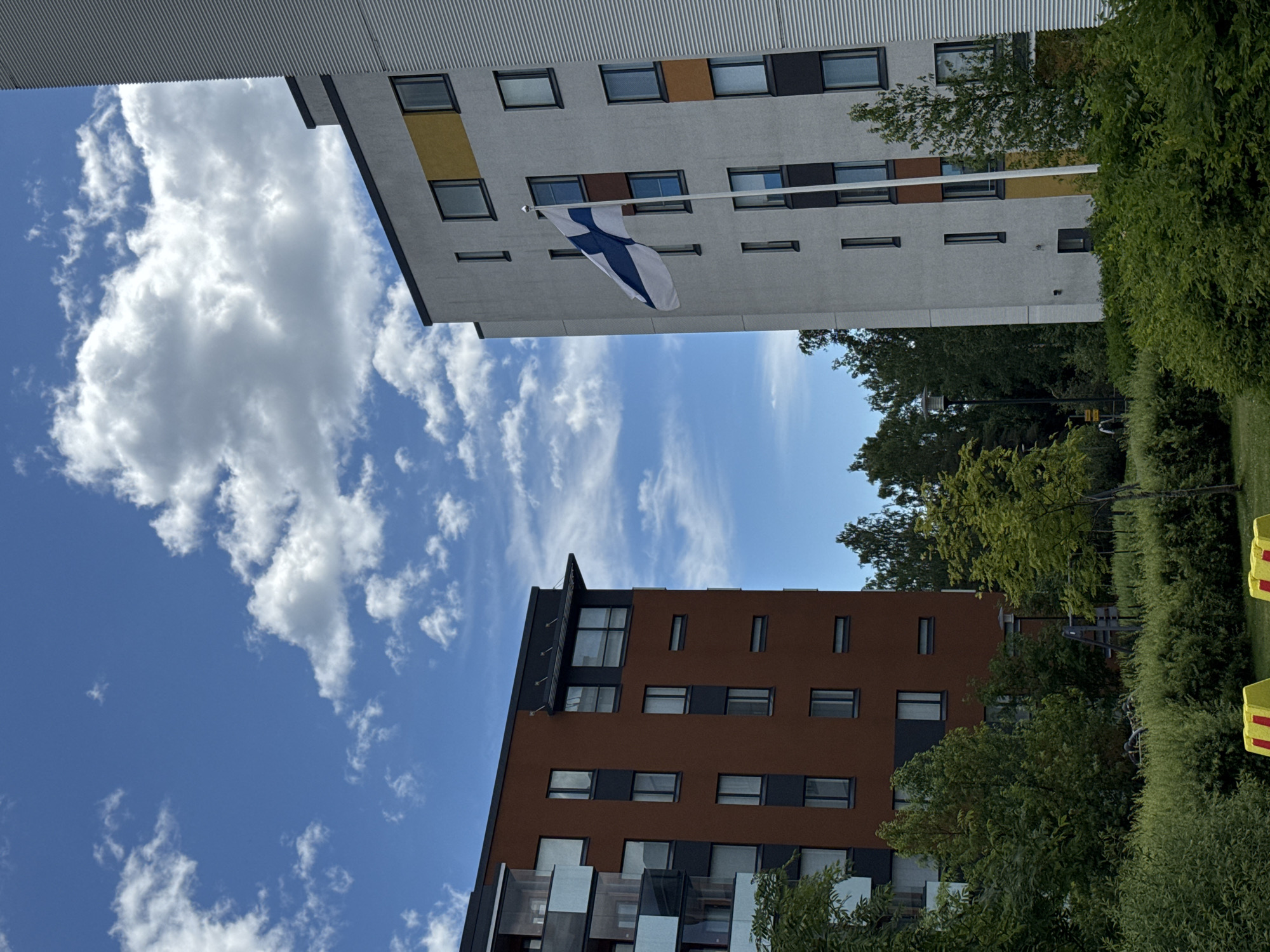 This media file is an outdoor, eye-level, vertical shot of two modern apartment buildings against a bright blue sky with scattered white clouds. The building in the foreground is clad in reddish-brown and dark gray panels, with numerous windows. The building in the background is lighter in color, with white and gray panels and accented with yellow and black sections. A Finnish flag is prominently displayed, hanging from a flagpole attached to the lighter building. The flag is white with a blue cross. Lush green trees and bushes flank the buildings, suggesting a park-like setting. The sun appears to be shining brightly, indicated by the clear sky and the way the light hits the buildings and foliage. There are some yellow, rectangular objects visible on the far right, possibly safety barriers or play equipment. The overall impression is one of a modern, well-maintained urban or suburban environment on a pleasant day. No people are visible in the image. There is no visible text in the image.