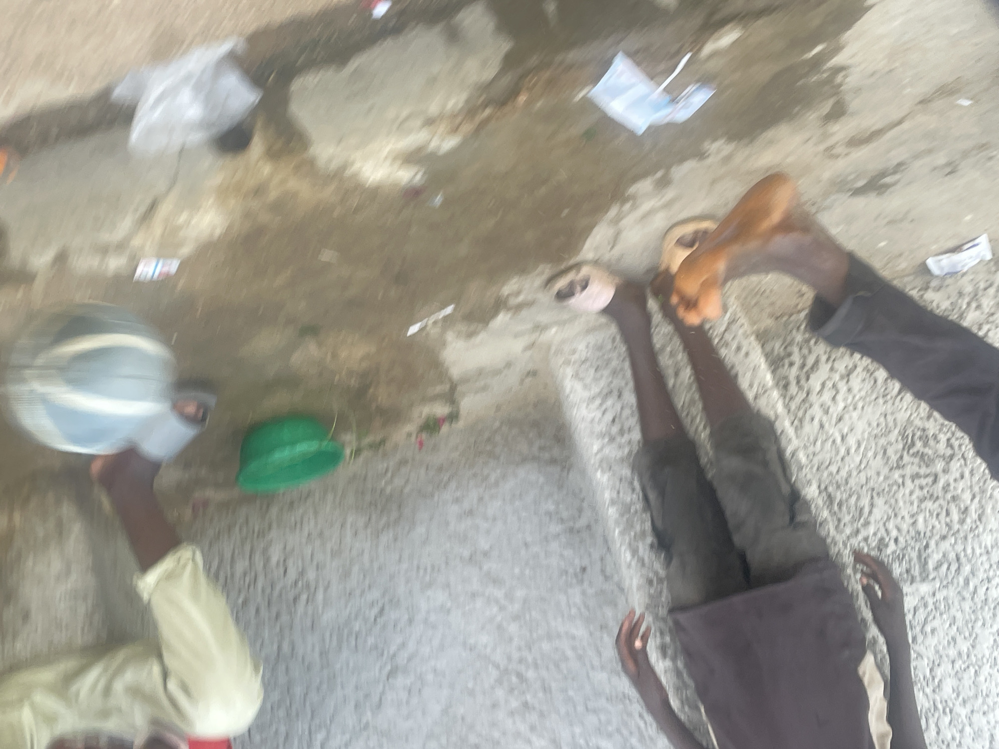 This overhead, blurry image captures an outdoor or semi-outdoor scene featuring at least two, possibly three, individuals on a concrete or tiled surface.
**Subjects:**
On the right side of the frame, a person's legs are visible, clad in dark trousers and wearing light-colored flip-flops or sandals. Their feet are bare. Further down, another person is seen lying down, with their hands and lower torso/legs in dark clothing resting on the rough concrete surface. On the left, an arm wearing a light-colored, long-sleeved garment holds a light-colored bowl or basin with a dark rim. Near this person, a green, possibly plastic, bowl or bucket is on the ground.
**Objects:**
Besides the clothing, footwear, and containers mentioned, the ground is littered with several small pieces of white paper or plastic, and a larger clear plastic bag is visible in the upper center.
**Scene and Setting:**
The setting appears to be a concrete area, possibly a courtyard, street, or a step-like structure. There are patches of what looks like water or dampness on the ground, creating dark stains and reflections. The texture of the ground varies, suggesting different surfaces or wear patterns. The overall environment seems informal and somewhat untidy due to the scattered litter.
**Activity or Event:**
The activity appears to be one of rest or informal gathering. One person is lying down, and another's legs are extended, suggesting relaxation. The person holding the bowl might be engaged in a domestic task like washing or preparing something. The blurriness of the image makes specific actions difficult to ascertain clearly.
**Notable Details:**
The image was taken during the daytime, indicated by the general light levels, although no direct sunlight is visible. The quality of the image is low, with significant blur, making fine details, facial expressions, and any visible text illegible. There are no clear cues regarding weather or a specific geographical location.