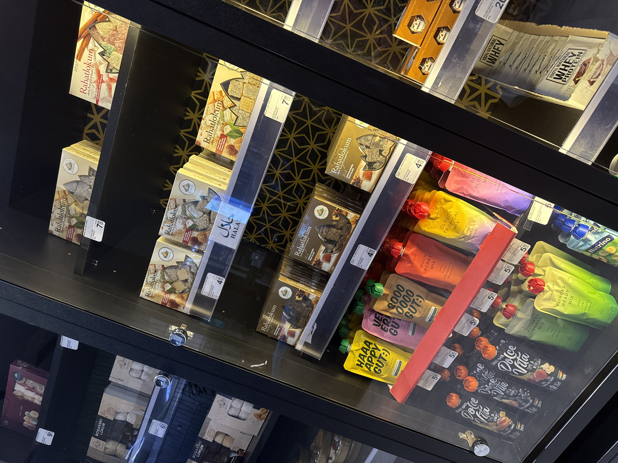 The image shows a refrigerated display case filled with food products, likely in a supermarket in Ilidža, Bosnia and Herzegovina. The products include various types of packaged sweets, notably "Rahatlokum" (Turkish delight), and pouches of nutritional drinks or mixes. Some of the products have Arabic script on their packaging, indicating an international selection. The display case is well-lit, and the glass front allows for a clear view of the merchandise. The overall impression is one of a clean, organized retail environment offering a diverse range of food items. There are no people visible in the image.