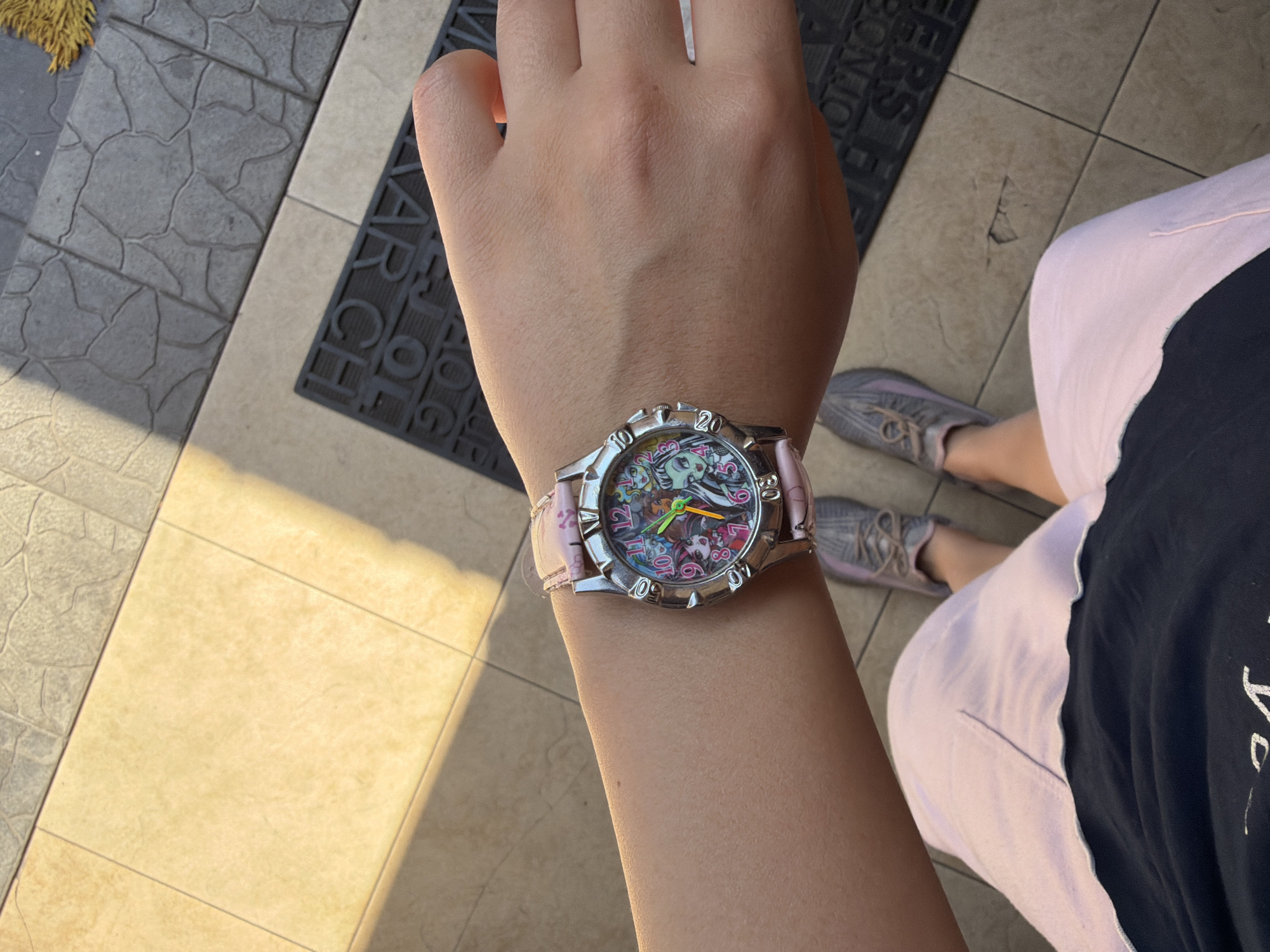 An overhead shot shows a person's right arm and wrist wearing a pink watch with a detailed face. The watch band is a light pink leather with a subtle pattern. The watch face features cartoon characters and colorful numbers. The person is wearing a dark t-shirt and light pink shorts or a skirt. The background consists of tiled flooring, with a darker mat visible near the bottom of the frame with embossed lettering. The lighting suggests an outdoor setting with sunlight casting shadows. The person's legs, wearing grey sneakers, are also visible in the background. There is no indication of the city of Qarağandı city administration, Kazakhstan in this image.