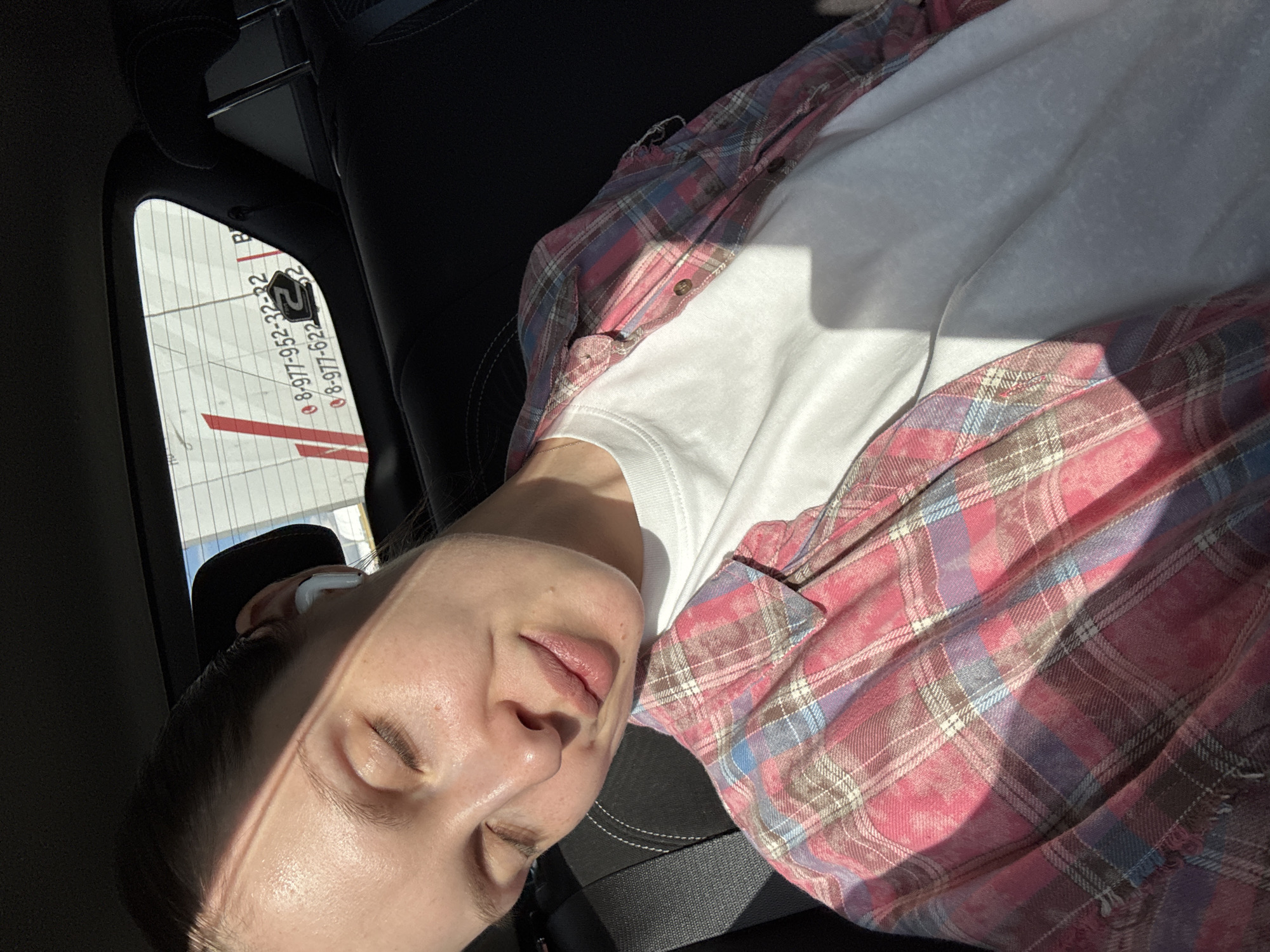 A pale-skinned female is reclining, possibly sleeping, in the back seat of a car. She is wearing a white T-shirt and a pink and blue plaid flannel shirt. She has what appears to be an earbud in her left ear. The sun is shining on her face. The car appears to be near an airport. Through the rear window, a building with numbers is visible: "8-977-952-32-32" and "8-977-622-62-62". The numbers suggest the scene may be in Russia, possibly Moscow based on the presence of a similar number format in that city. The exact location within Moscow cannot be determined from the information.
