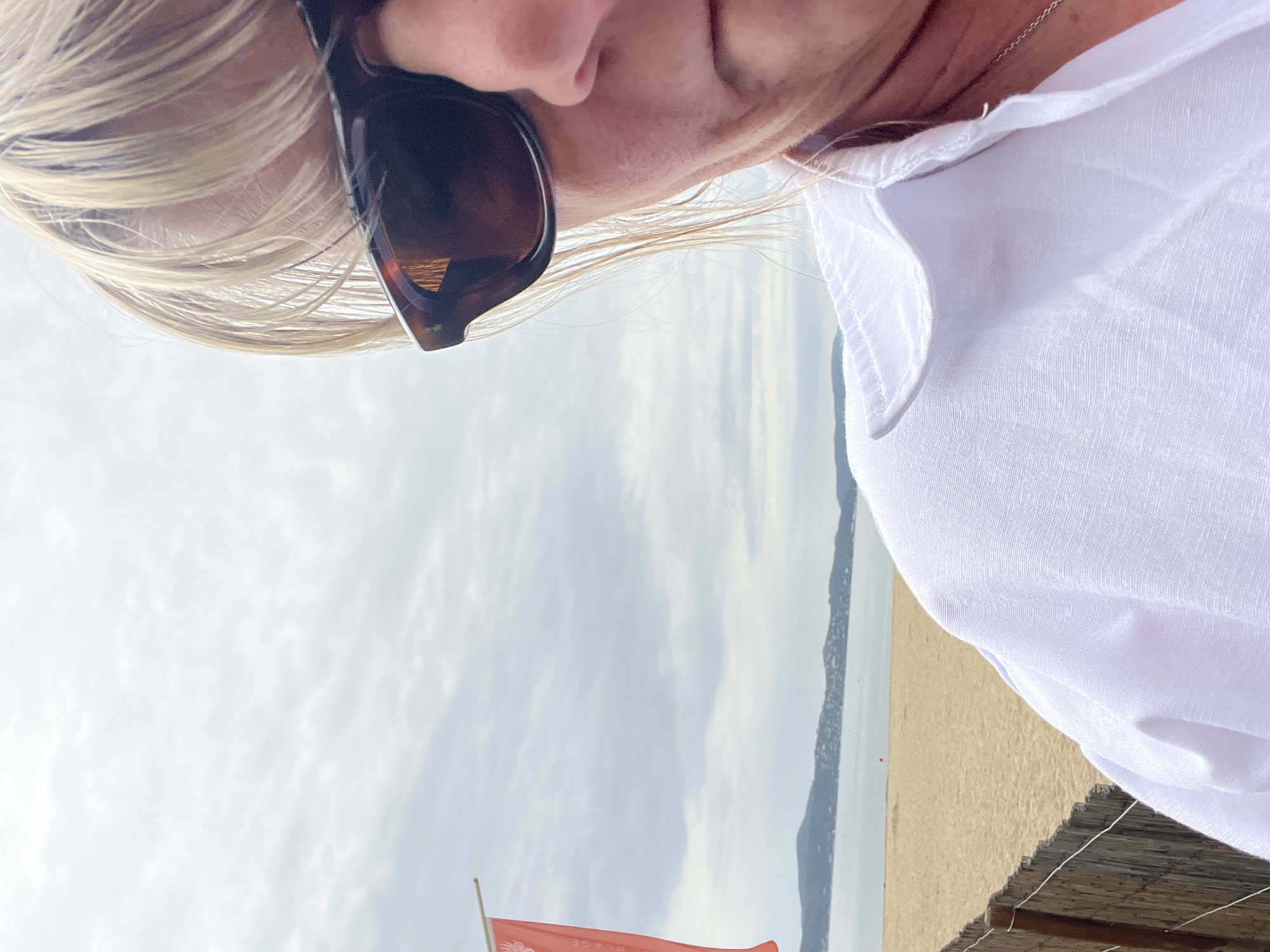 A close-up, candid photograph captures a person with blonde hair and sunglasses at a beachside location in Sainte-Maxime, France. The person is wearing a white shirt. The photo appears to be taken from a slightly downward angle. The background includes a beach, ocean, and a cloudy sky. A red flag and a thatched roof structure are also visible.