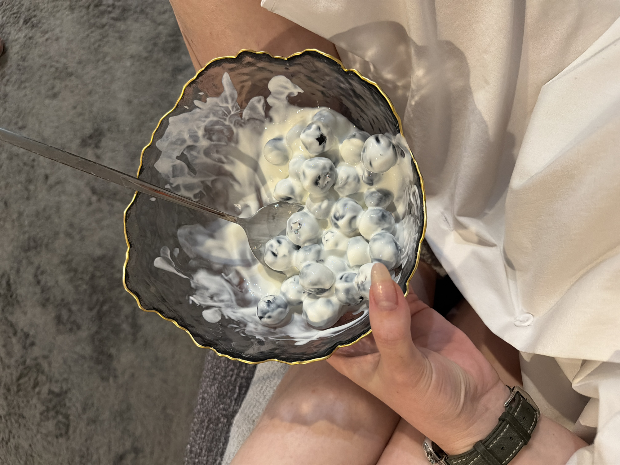 An overhead, close-up shot features a person holding a decorative bowl filled with blueberries generously coated in a white, creamy substance, likely yogurt or cream. The bowl itself is a notable object, with an irregular, wavy rim edged in gold, and appears to be crafted from dark, possibly translucent, glass or ceramic, featuring subtle white marbling patterns within its material. A silver spoon is partially submerged in the creamy mixture, indicating the person is either preparing to eat or is in the process of consuming the snack.
Visible parts of the person include their bare legs, with one knee slightly prominent, and their right hand holding the bowl. Their left wrist, sporting a watch with a dark green or gray leather strap and a silver buckle, is also visible. The person is wearing a light-colored, possibly white or off-white, soft fabric garment, draped around their lap and extending into the background on the right side of the frame.
The setting appears to be indoors, with a dark gray, textured carpet or rug visible in the background on the left. The lighting is even, but no specific time of day or outdoor weather conditions can be discerned. There is no visible text in the image. The scene conveys a moment of casual enjoyment or a snack break.