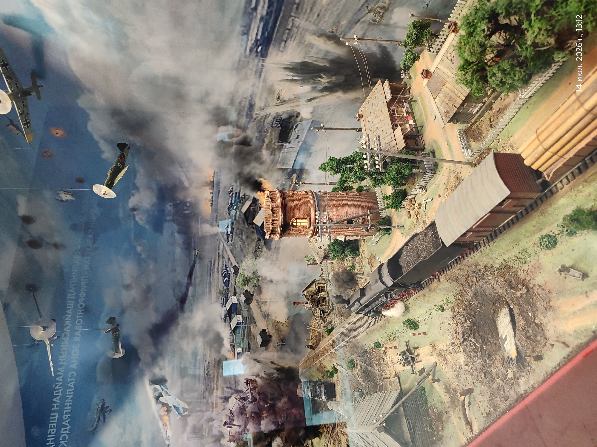 This is a diorama depicting a wartime scene, likely a diorama at a museum. The diorama features a miniature village or town under attack, with buildings in ruins, smoke billowing from explosions, and a train on tracks. Several model airplanes are suspended in the air, some appearing to be in combat. The overall scene conveys destruction and conflict. There is text in Russian at the bottom right corner that reads "14 июля 2026 г., 13:12", which translates to "July 14, 2026, 13:12", indicating the date and time the photograph was taken, not the setting of the diorama. There is no information within the image that specifies the location as Astana, Kazakhstan, or any other specific city.