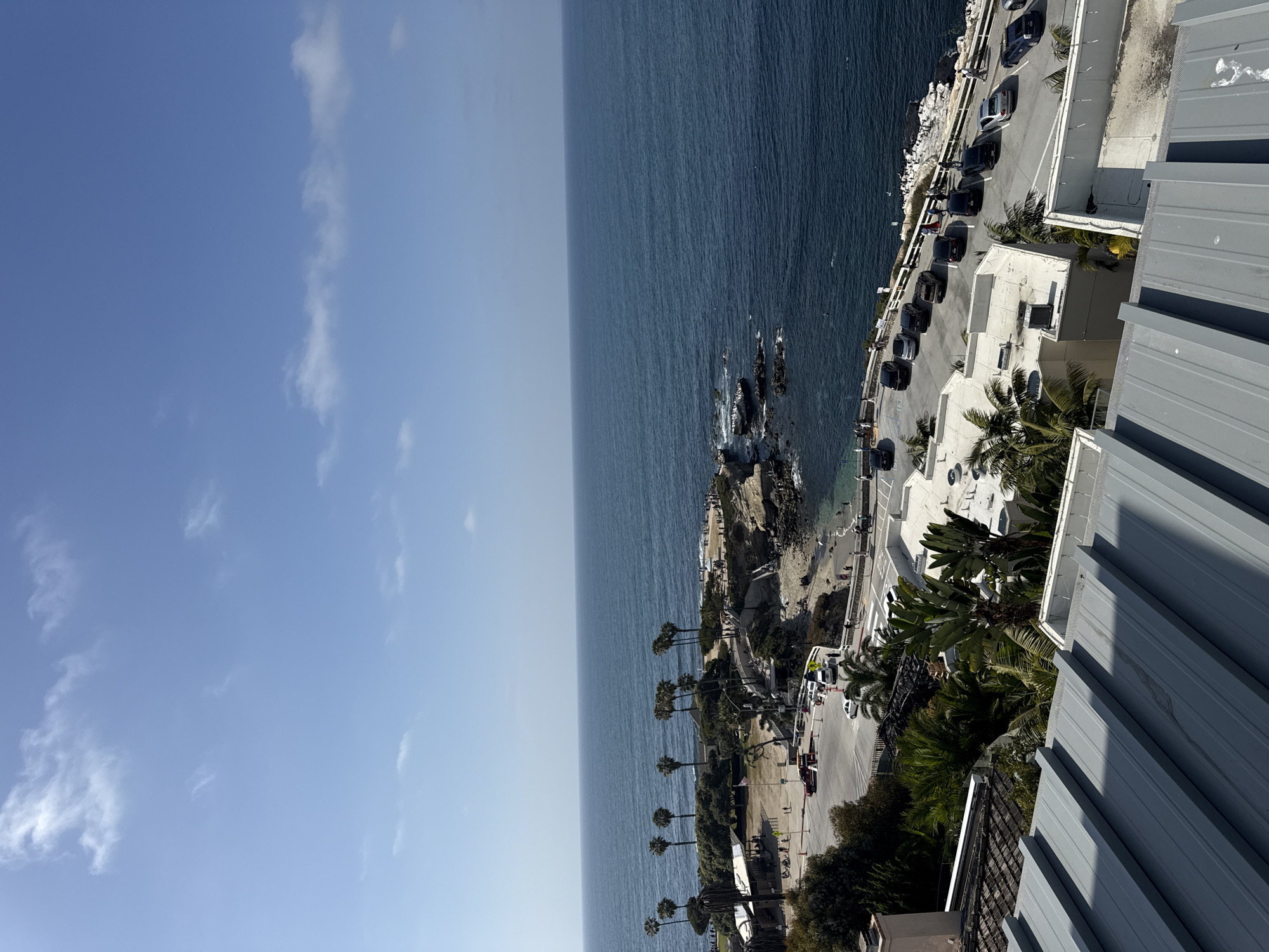 The image shows an aerial view of the coastline in San Diego, United States of America. The scene is dominated by the ocean, which stretches from the center to the left of the frame and meets a clear, blue sky with scattered clouds. A rocky coastline with cliffs extends from the ocean's edge towards the right side of the image. A road runs along the top of the cliffs, lined with parked cars. A building with a corrugated metal roof occupies the bottom right corner of the frame, suggesting that the photograph was taken from an elevated position within or near the structure. Palm trees and other vegetation are visible along the road and near the building. Small figures of people can be seen walking along the path at the cliff's edge.