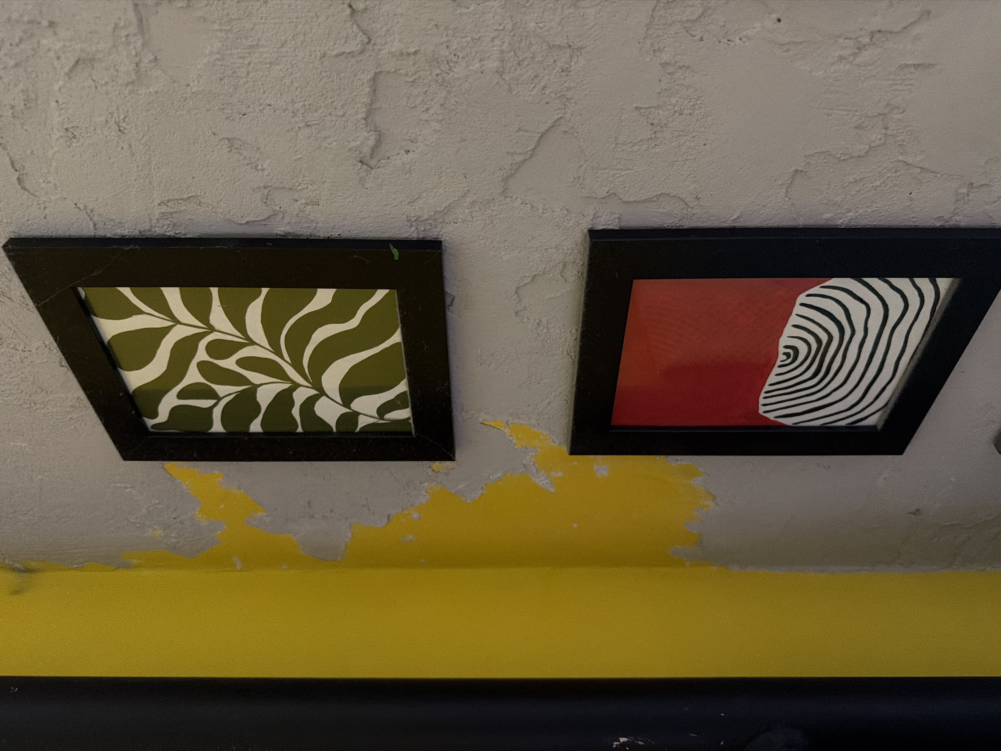 The image shows a corner of a room with a textured grey wall and a vibrant yellow painted column. Two framed pictures are hung on the grey wall. The top picture features a botanical print with green wavy leaf shapes on a white background. The bottom picture has a solid red background with a black and white abstract pattern of concentric curved lines. The yellow paint on the column appears to be peeling in places, revealing the grey wall underneath. A dark vertical object, possibly a door frame or pillar, is visible on the far left edge of the image. The lighting suggests an indoor setting, possibly a home or a public space like a cafe or gallery. The location is not specified in the image.