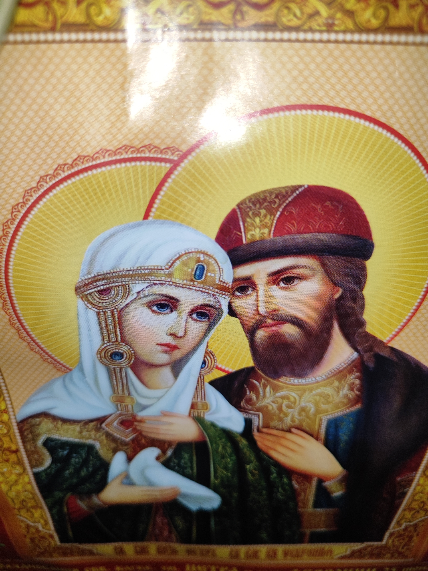 This is a close-up, eye-level, indoor photograph of an icon, likely religious in nature. The icon depicts two figures, a man and a woman, who appear to be saints or biblical figures. The woman is on the left, facing slightly away from the viewer, with a white veil and an ornate headdress adorned with jewels. She has blue eyes and is holding a white dove in her hands. The man is on the right, facing more towards the viewer, with a beard, wearing a red and gold hat and richly decorated clothing. Both figures have halos behind their heads, depicted as yellow, radiating circles. The background is a patterned yellow and gold fabric. The overall style is typical of Eastern Orthodox iconography. The image quality is good, with clear details and vibrant colors. The lighting suggests it is illuminated by an artificial light source, possibly a flash, which creates a glare on the surface of the icon. There is no indication of the specific location within Fergana City, Uzbekistan, as the image focuses solely on the icon itself.