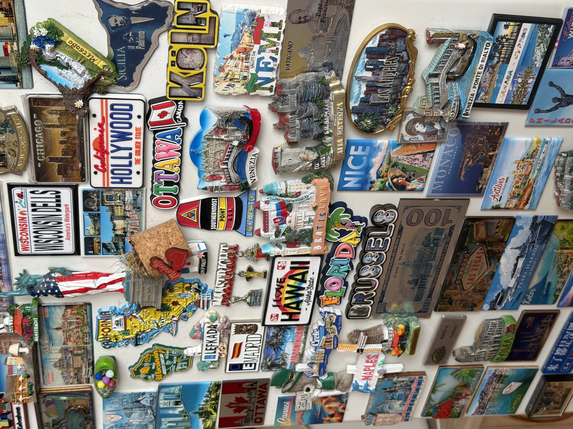 The image shows a collection of magnets attached to a white surface, likely a refrigerator. The magnets are souvenirs from various locations around the world, including cities, states, and countries. Locations include:
- Europe: Venice, Rome, Brussels, Naples, Crete, Cyprus, Monaco, Nice, Madrid, Bratislava, Ohrid, Nemi
- United States: New York, Chicago, Wisconsin, California, Hawaii, Florida, Las Vegas, Pearl Harbor
- Canada: Ottawa
- Other: Sicily, Vatican City, Istanbul
No people, subjects, actions, interactions, or location in the city of Buhovo, Bulgaria are evident in the image.