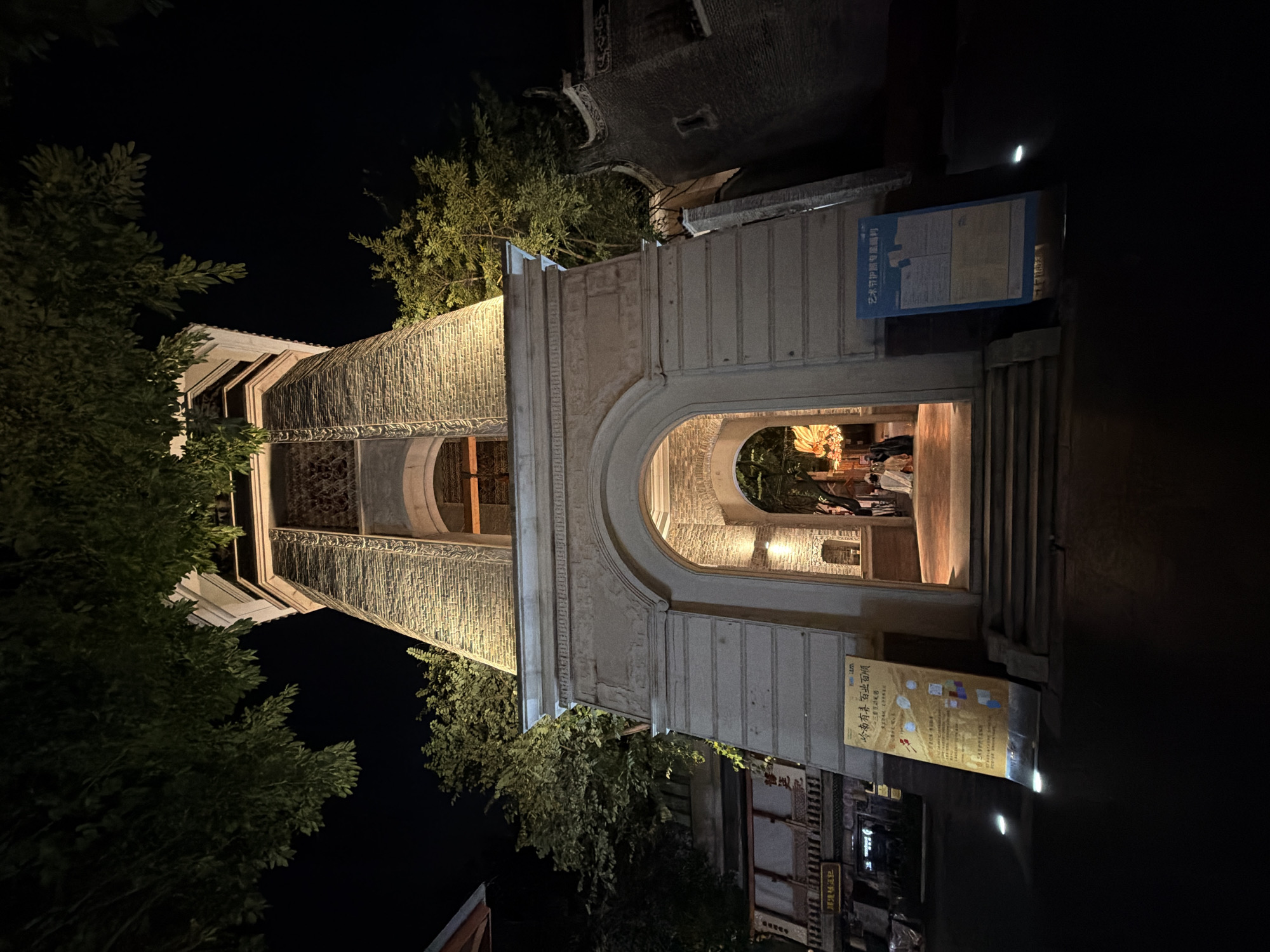 A wide-angle, low-angle nighttime exterior shot captures a brick tower structure with an arched entrance in Chancheng District, China. The tower, made of textured bricks, is illuminated from below, highlighting its architectural details and casting light upwards. Above the archway, decorative elements are visible. Lush green trees overhang the top of the scene, obscuring a portion of the sky and the upper parts of the tower. To the left of the tower, a multi-story building with a dark facade is partially visible, adorned with signage. On the right side of the entrance, a smaller, darker brick building with a curved roofline is attached to the tower. Inside the arched entrance, a warmly lit walkway leads to an interior space where several people are gathered, one person in a white garment and others in darker clothing. The ground in front of the tower is paved and reflects ambient light. Two upright display boards are positioned near the entrance; one is yellow with Chinese text and graphics, and the other is blue with Chinese text, both illuminated by spotlights. The overall atmosphere is a mix of historic architecture and evening activity.