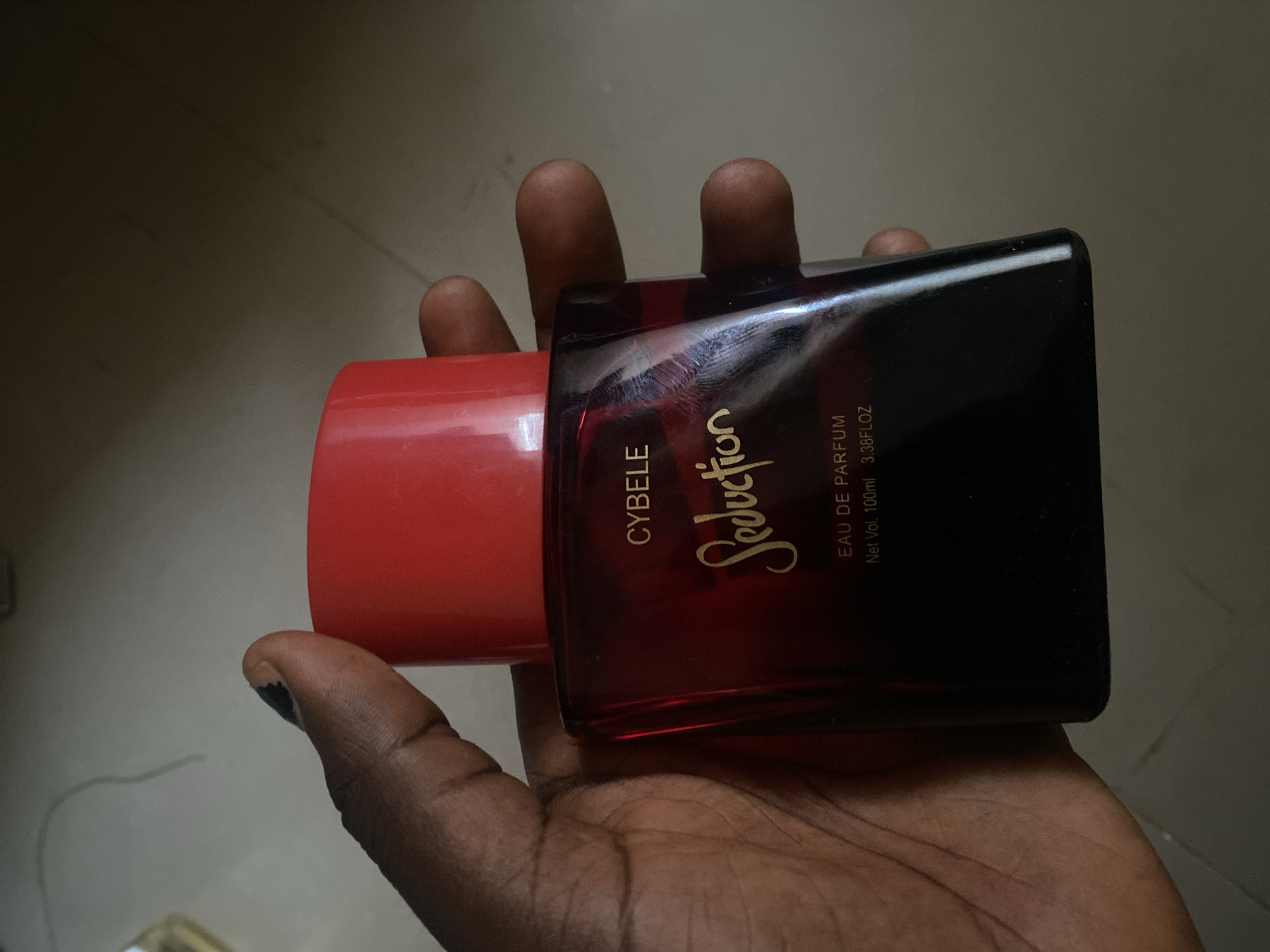 A hand holds a bottle of "Seduction" by Cybele perfume. The bottle is dark red with a gradient to black, and the cap is bright red. Text on the bottle reads "CYBELE," "Seduction," "EAU DE PARFUM," and "Net Vol. 100ml 3.38FLOZ." The hand holding the bottle appears to be that of a person with dark skin, with dark nail polish visible on one of the fingers. The background is out of focus, showing a muted, neutral-colored surface. The lighting suggests an indoor setting, possibly during the daytime. The image is cropped to focus on the hand and the perfume bottle.