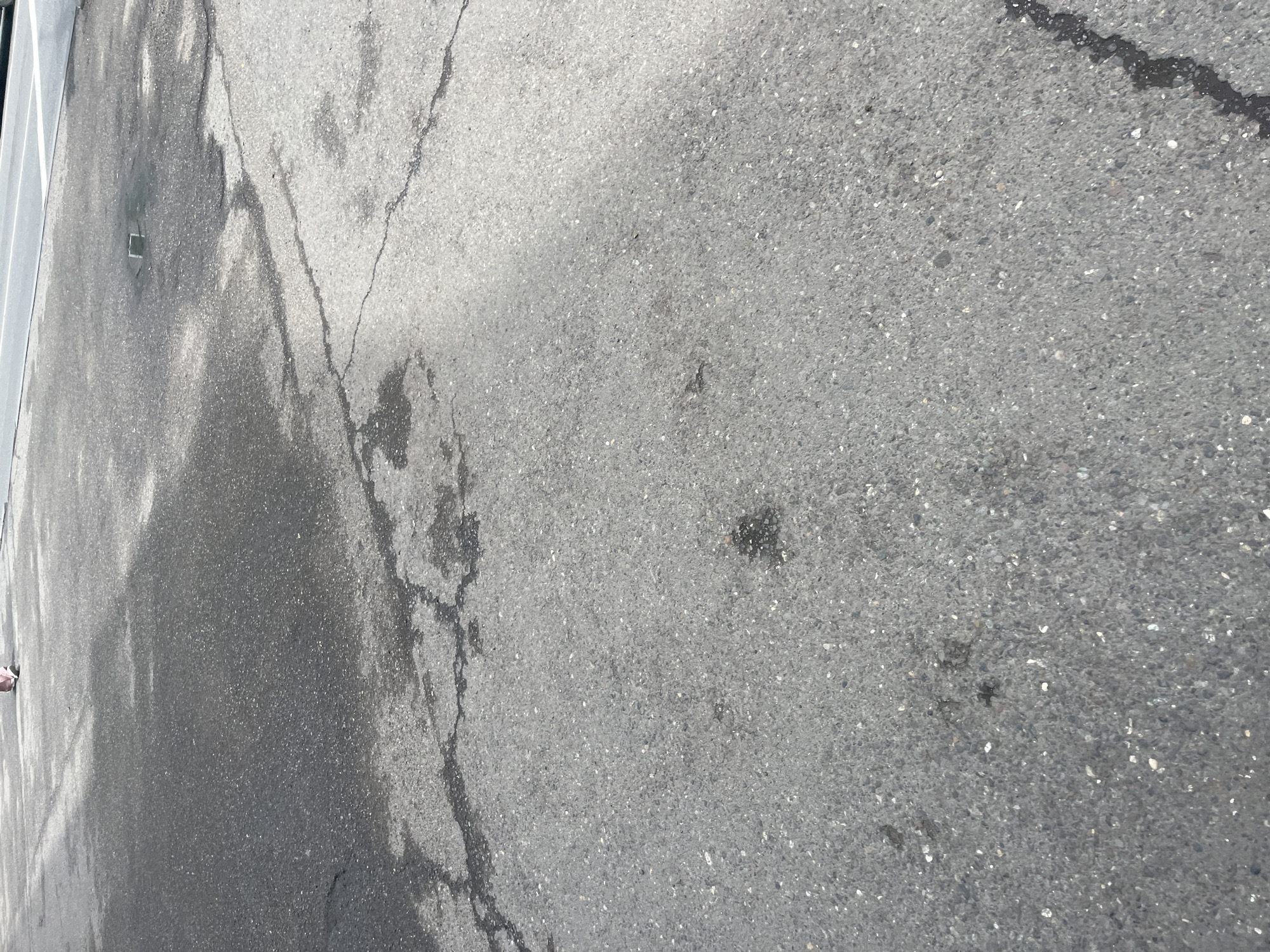 The image depicts cracked asphalt pavement. The pavement is gray, with varying degrees of wear and discoloration. There are several distinct cracks running across the surface. The location is not discernible from the image. No people, subjects, objects, actions, or interactions are visible.